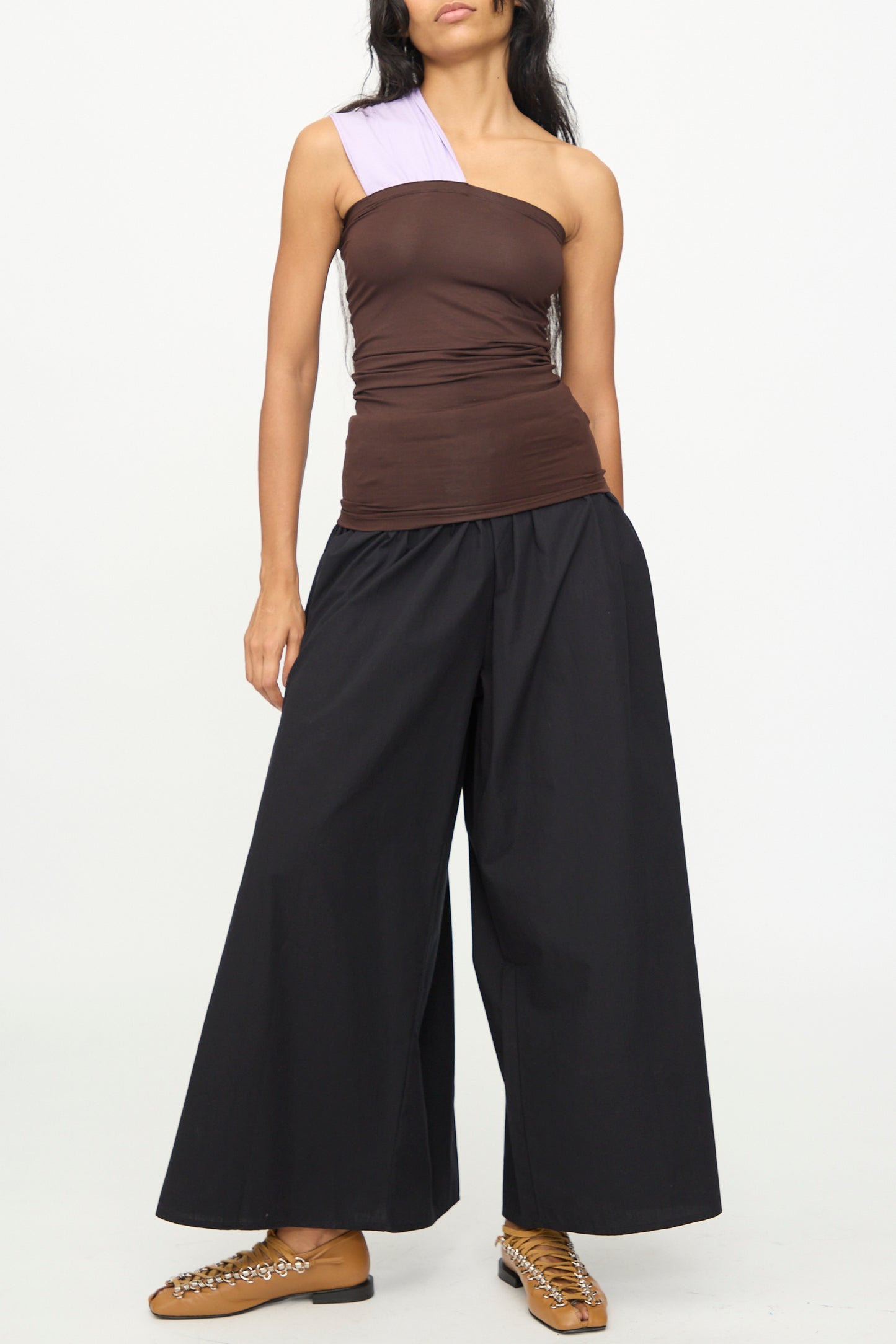 A woman wears the Baserange Geo Tank in Tactile One Purple, featuring a brown one-shoulder design with a light purple single shoulder strap, paired with black wide-leg pants and tan shoes with chain details, standing against a plain white background.