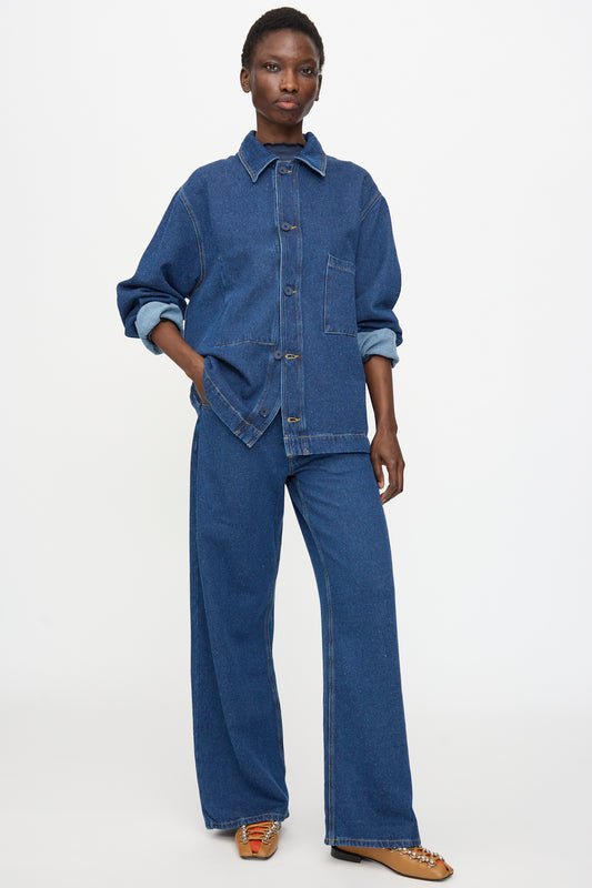 A person stands against a plain background wearing Baserange Grass Pants in Indigo Blue Wash, styled with a matching denim jacket and brown flat shoes.