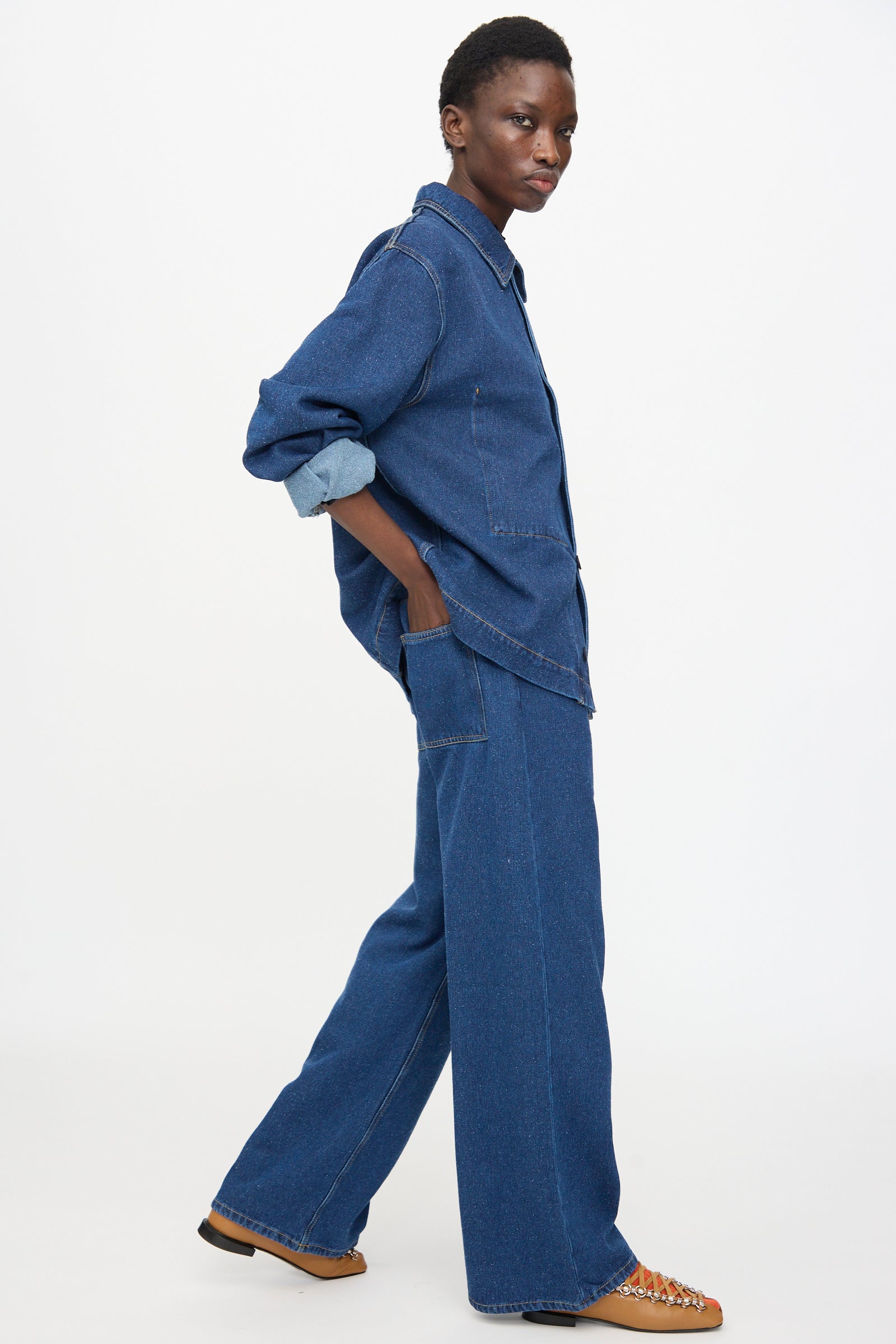 A person stands sideways against a plain white backdrop, hands in pockets, wearing Baserange Grass Pants in Indigo Blue Wash with a matching loose denim shirt and brown lace-up shoes.