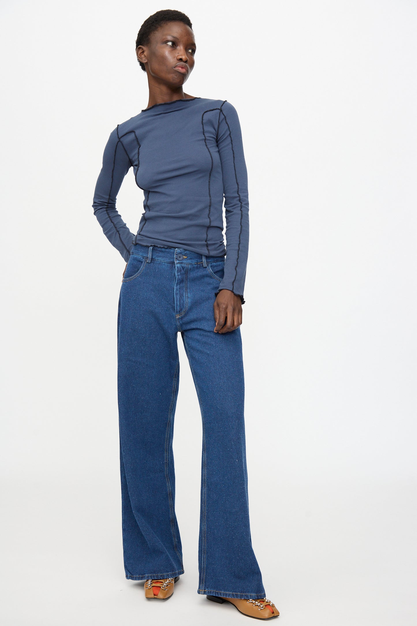 Person stands against a plain background, wearing a long-sleeve blue top, Baserange Grass Pants in Indigo Blue Wash, and brown shoes.