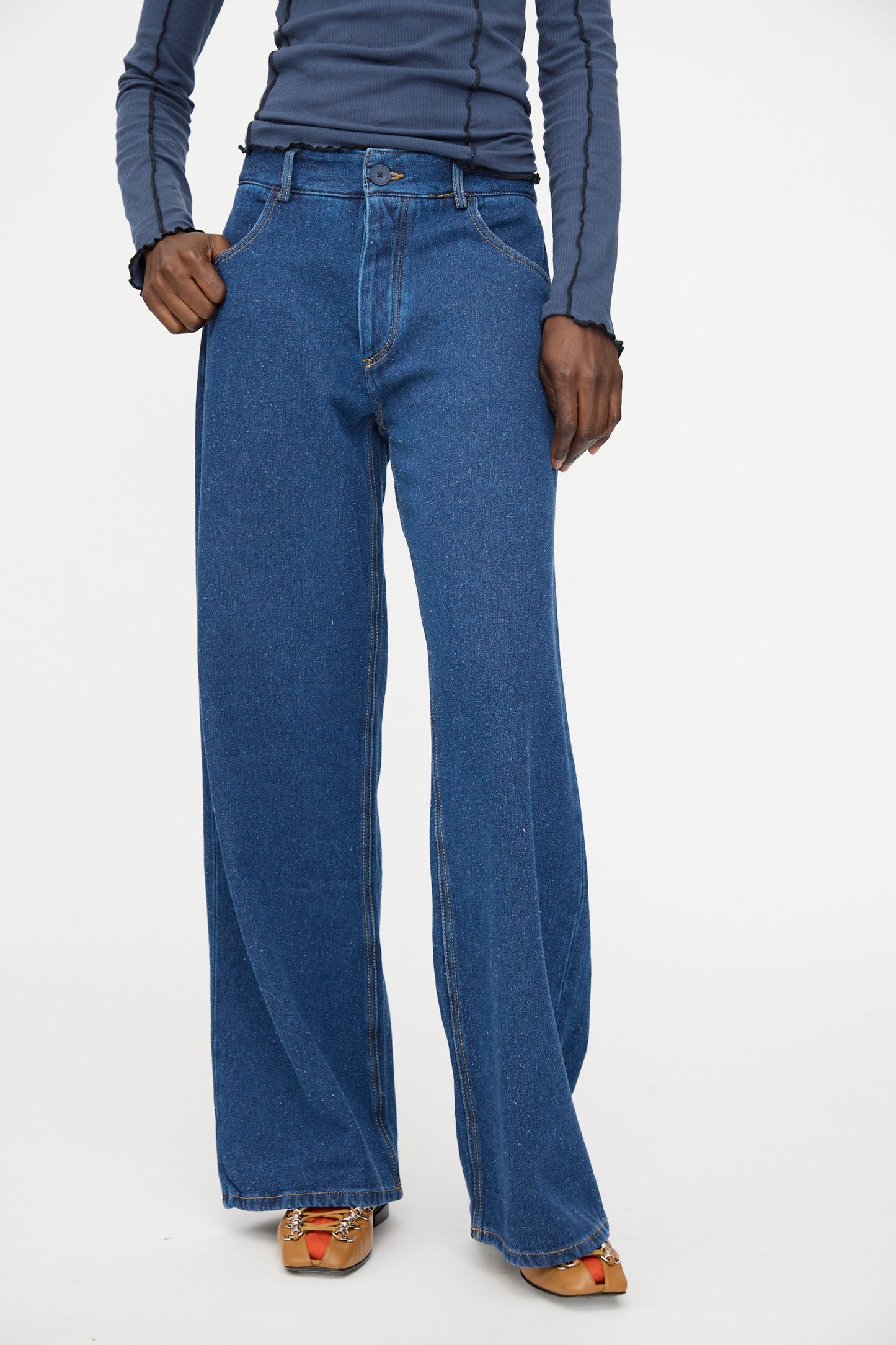A person wearing Baserange Grass Pants in Indigo Blue Wash, a long-sleeve indigo blue silk-cotton blend top, and open-toed shoes stands against a plain white background.