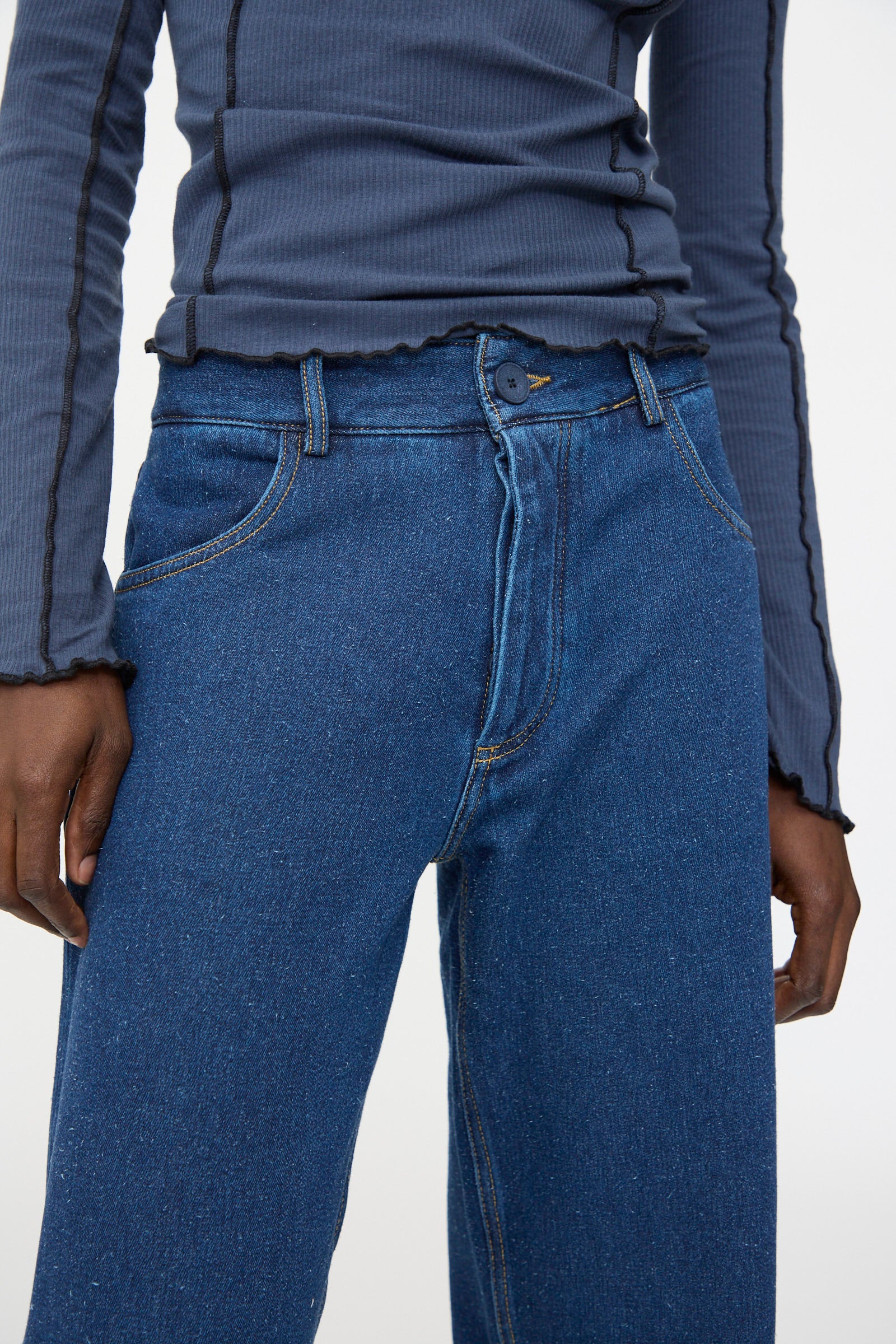 A person wearing a ribbed, long-sleeve dark gray top and Baserange Grass Pants in Indigo Blue Wash, standing with arms relaxed at their sides.