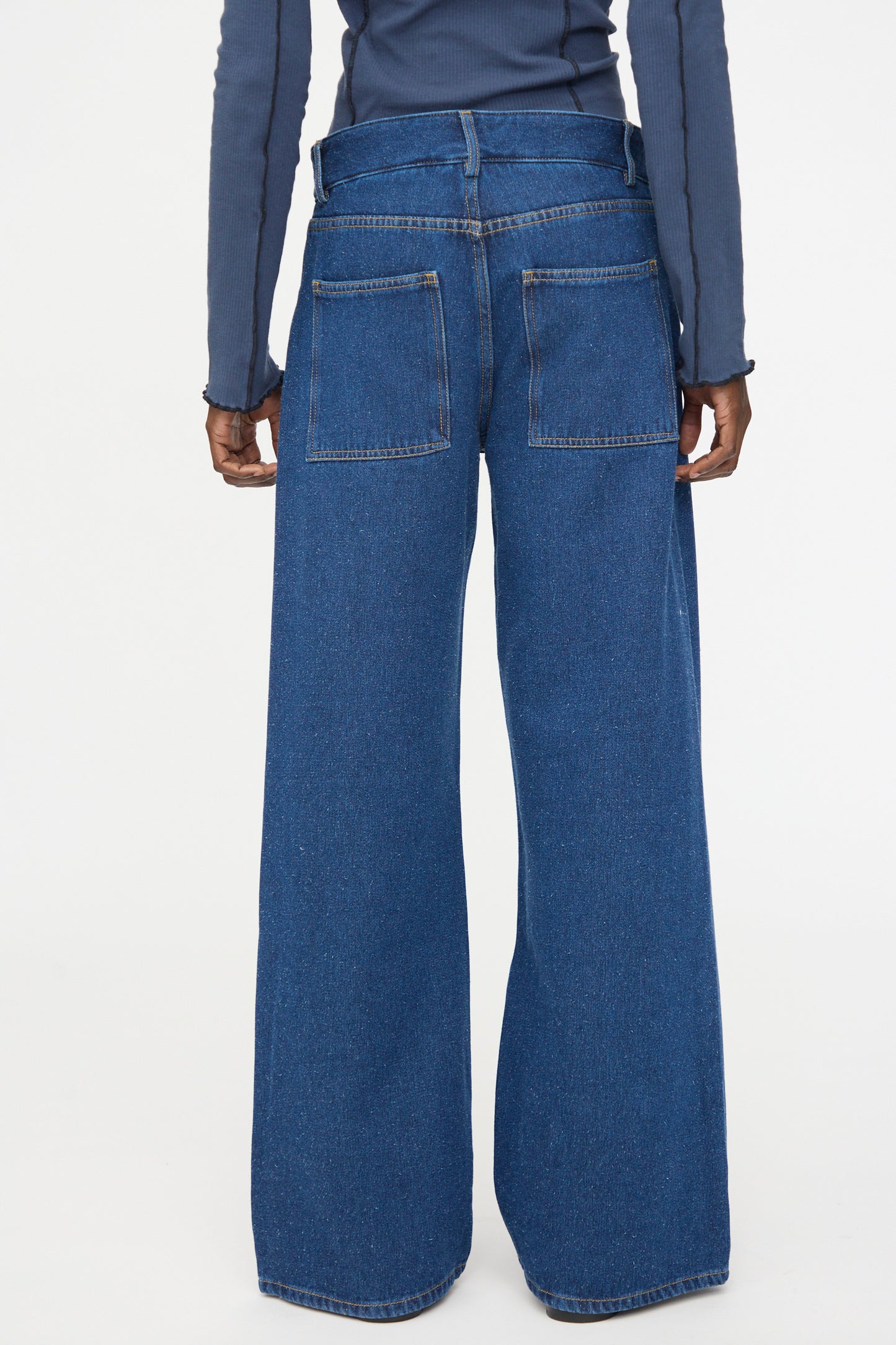 A person stands facing away, wearing Baserange Grass Pants in Indigo Blue Wash—wide-leg jeans with a double waistband and back pockets, crafted from a silk-cotton blend—paired with a long-sleeve blue shirt.