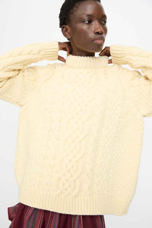 A person wearing the Lens Sweater in Chaume Sand by Baserange and a burgundy skirt stands against a plain white background, adjusting the collar of the sweater.