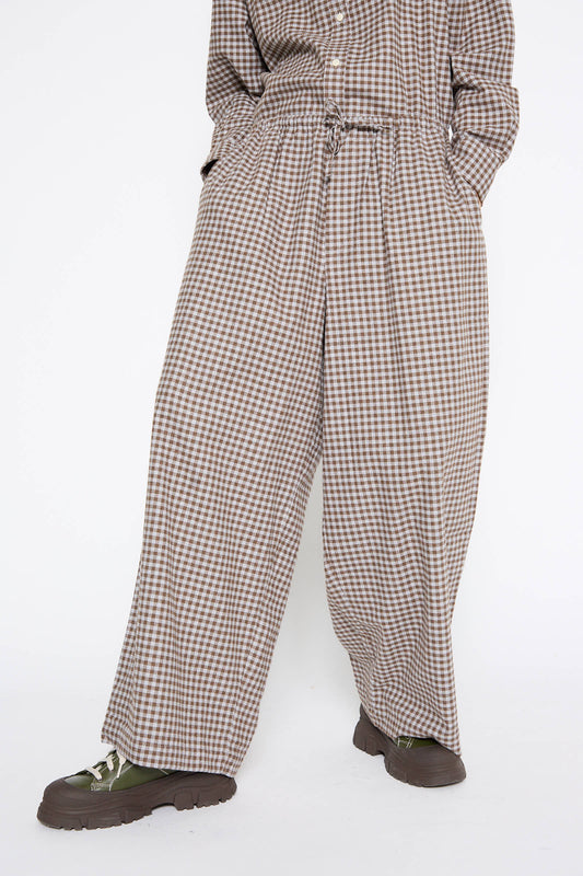 Person wearing a matching checkered outfit with wide-leg, relaxed fit Limber Pant in Brown Grey Flannel by Baserange, featuring an elasticated waist, paired with a button-up check flannel shirt and green lace-up shoes.