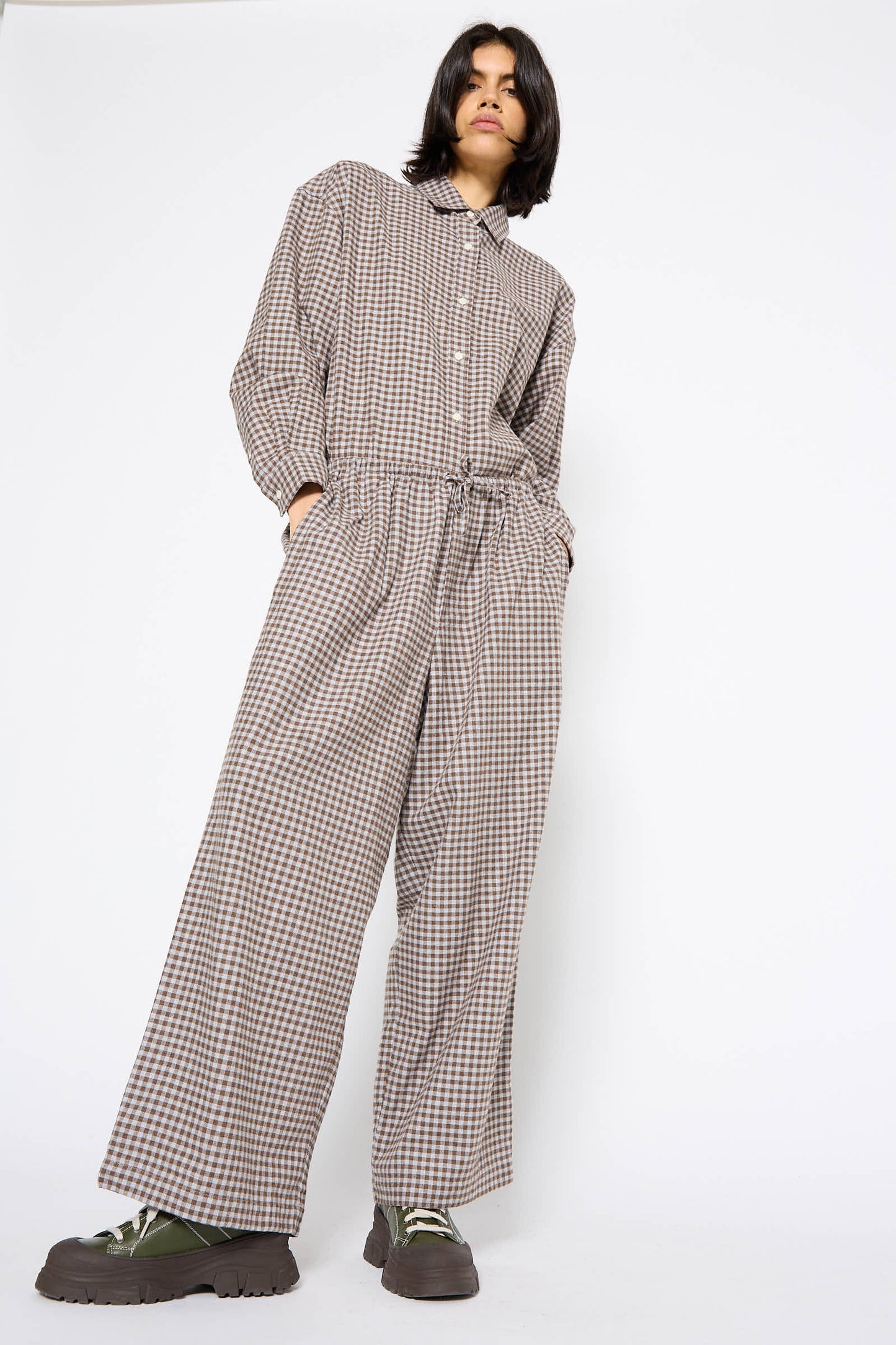 A person stands in a loose-fitting checkered jumpsuit with an elasticated waist, hands in pockets, looking slightly downwards. The outfit includes the Limber Pant in Brown Grey Flannel from Baserange and large brown platform shoes with green laces.