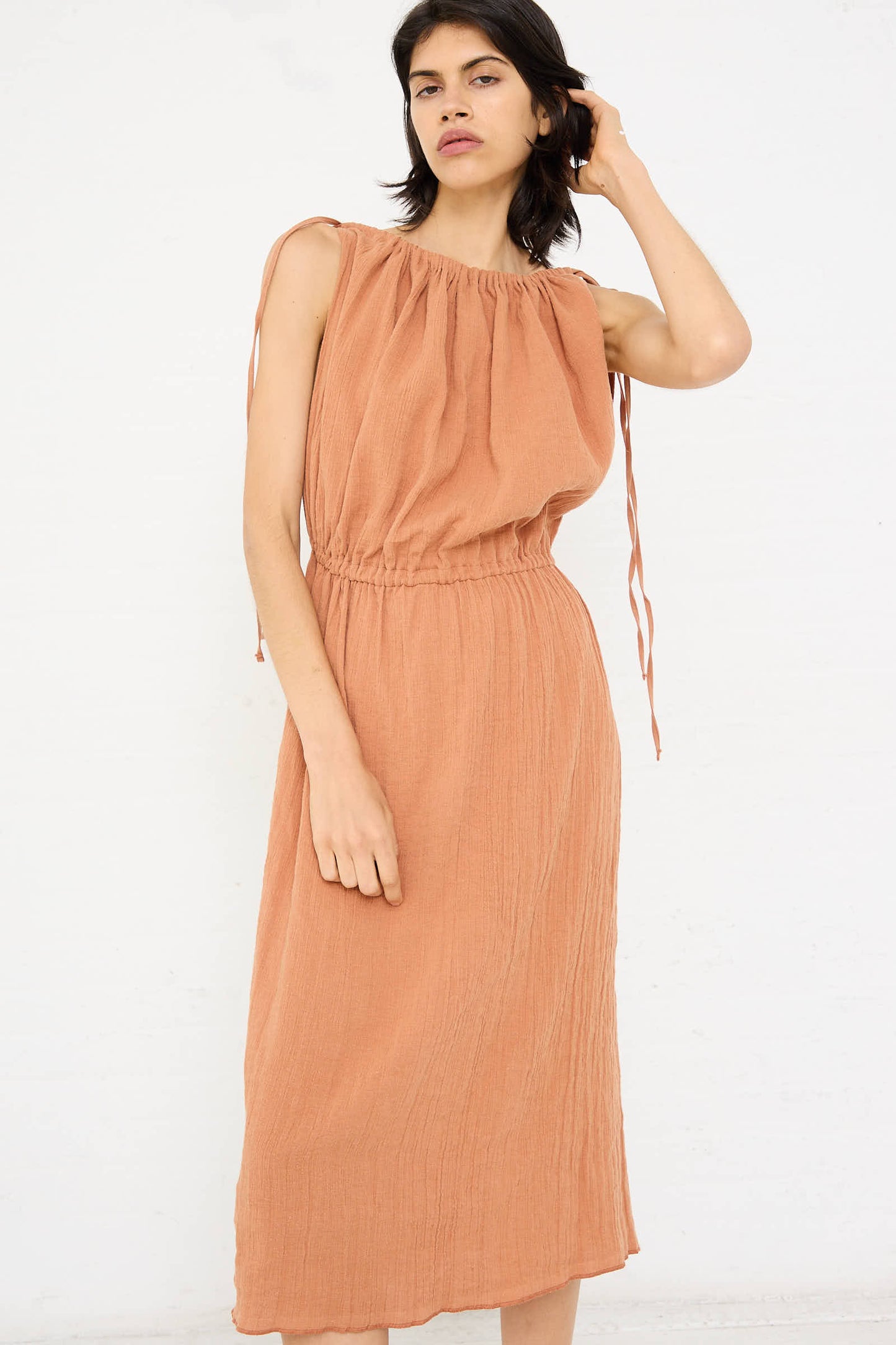 A person wearing a sleeveless Brick Linen Cotton Diph Dress with a gathered neckline and waist by Baserange, standing against a plain white background.