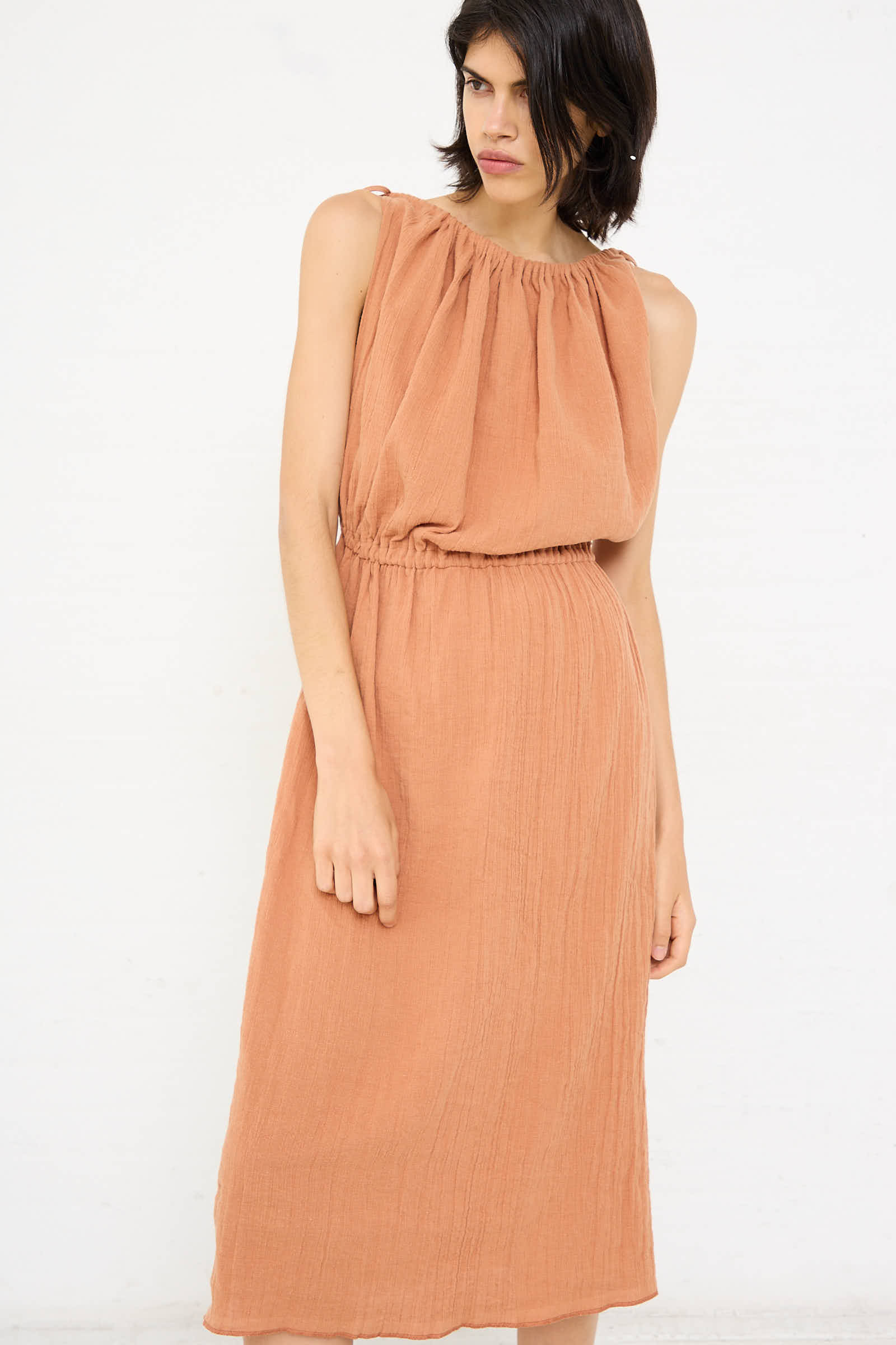 A person is looking down, wearing the Baserange Linen Cotton Diph Dress in Brick, a sleeveless, calf-length dress crafted from crinkled linen with an elastic waist and a gathered neckline. They stand against a white background.