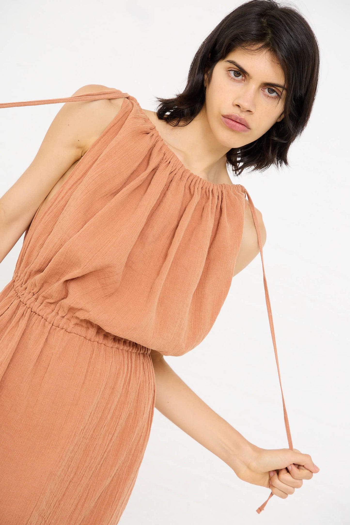 A person wearing the Linen Cotton Diph Dress in Brick by Baserange, featuring sleeveless design with gathered shoulder details and a relaxed fit, looks towards the camera against a white background.