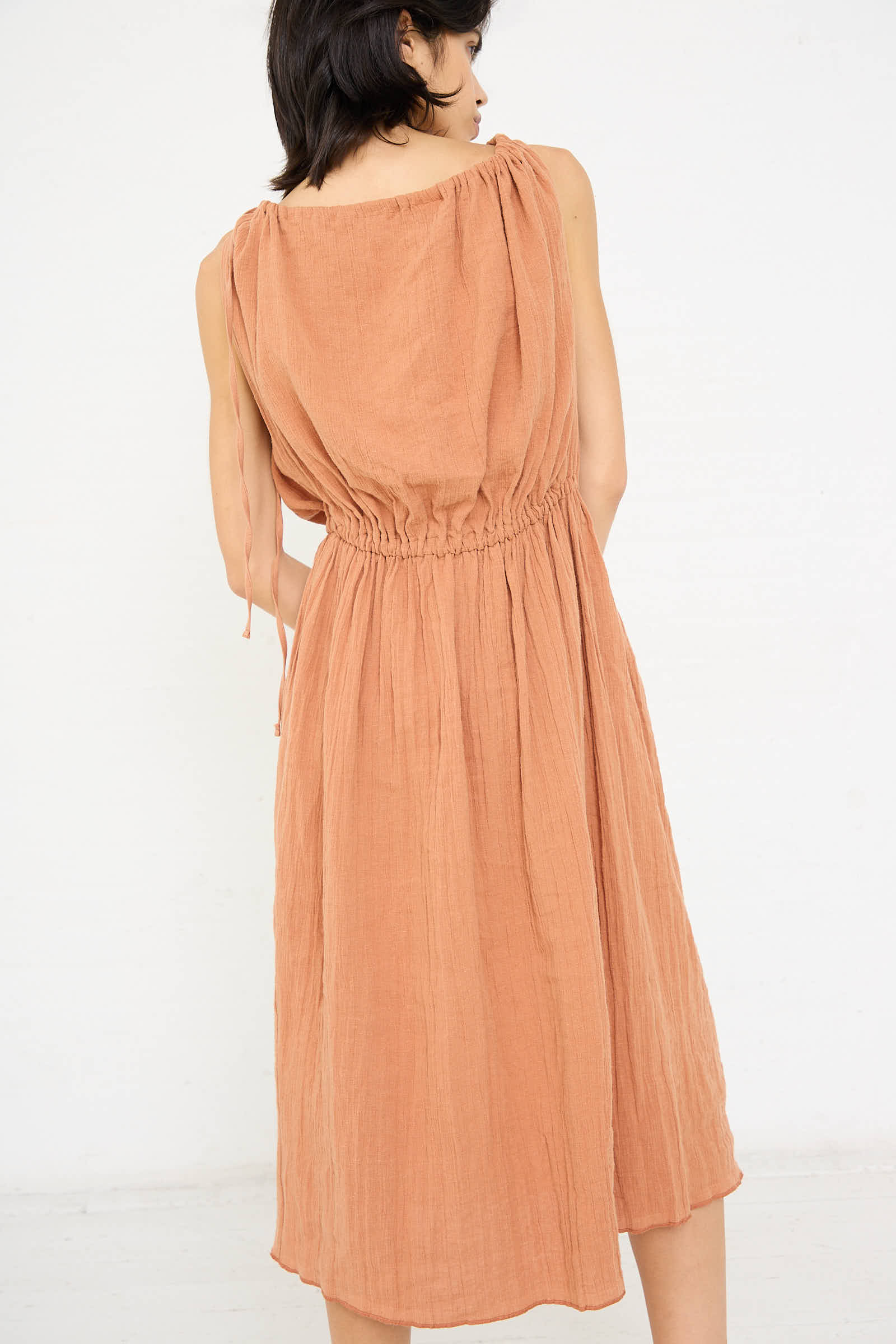 A person with short dark hair is facing away from the camera, wearing the Linen Cotton Diph Dress in Brick by Baserange, which features a sleeveless design, a knee-length cut, a gathered waist, and a relaxed fit.