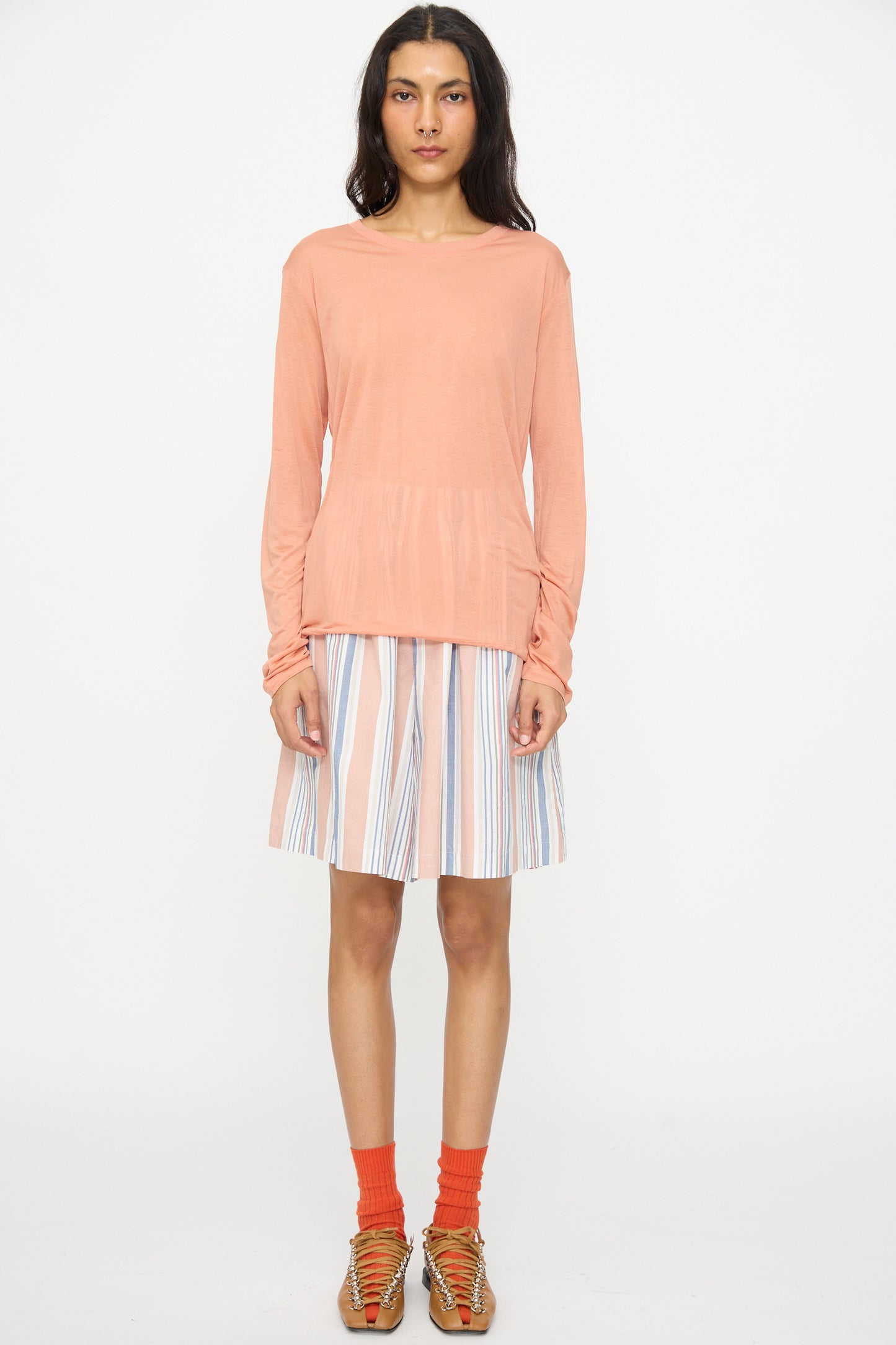 A person stands facing forward against a plain white background, wearing the Baserange Long Sleeve Tee in Branch Terracotta, a striped knee-length skirt, orange socks, and brown woven sandals.