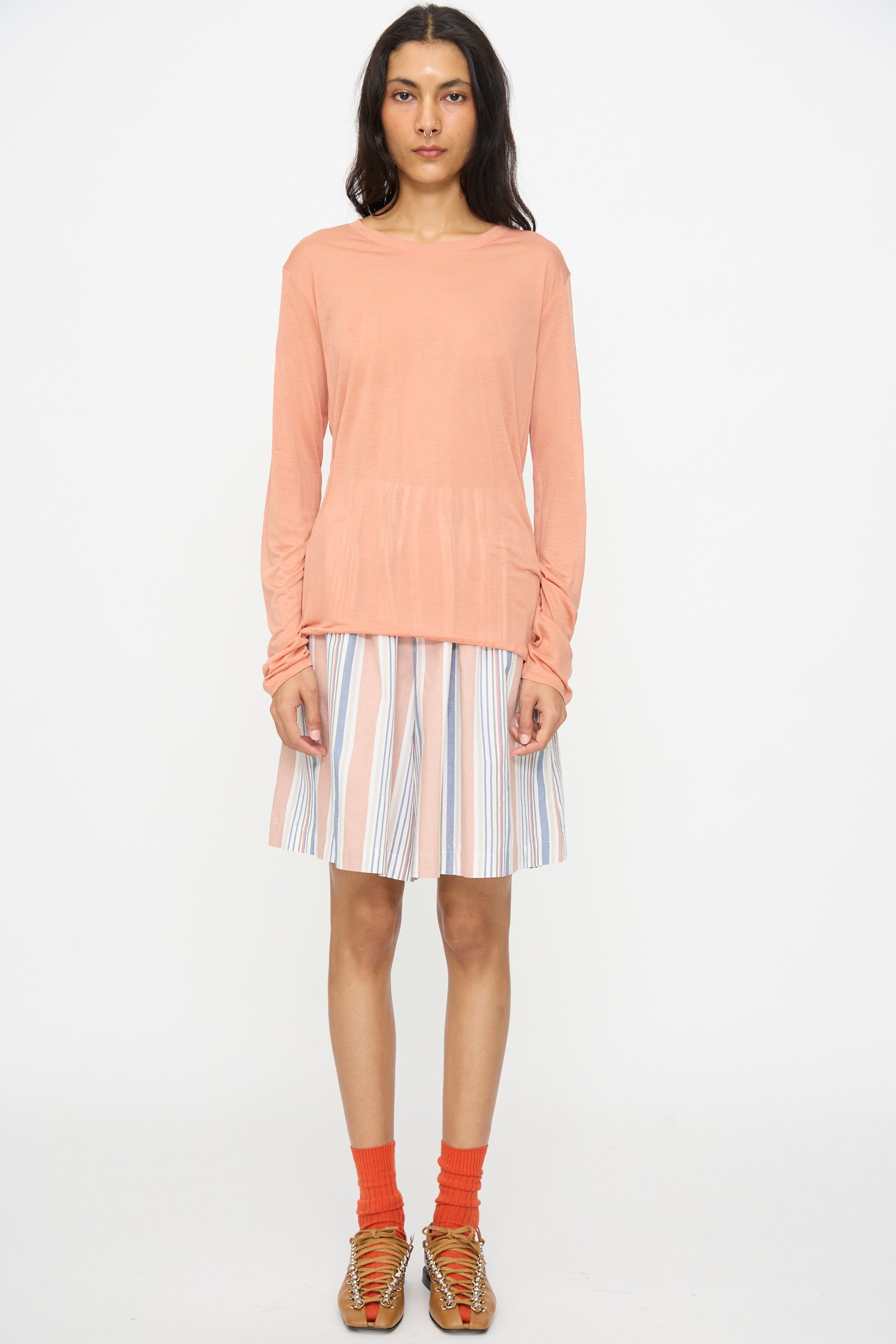 A person stands facing forward against a plain white background, wearing the Baserange Long Sleeve Tee in Branch Terracotta, a striped knee-length skirt, orange socks, and brown woven sandals.