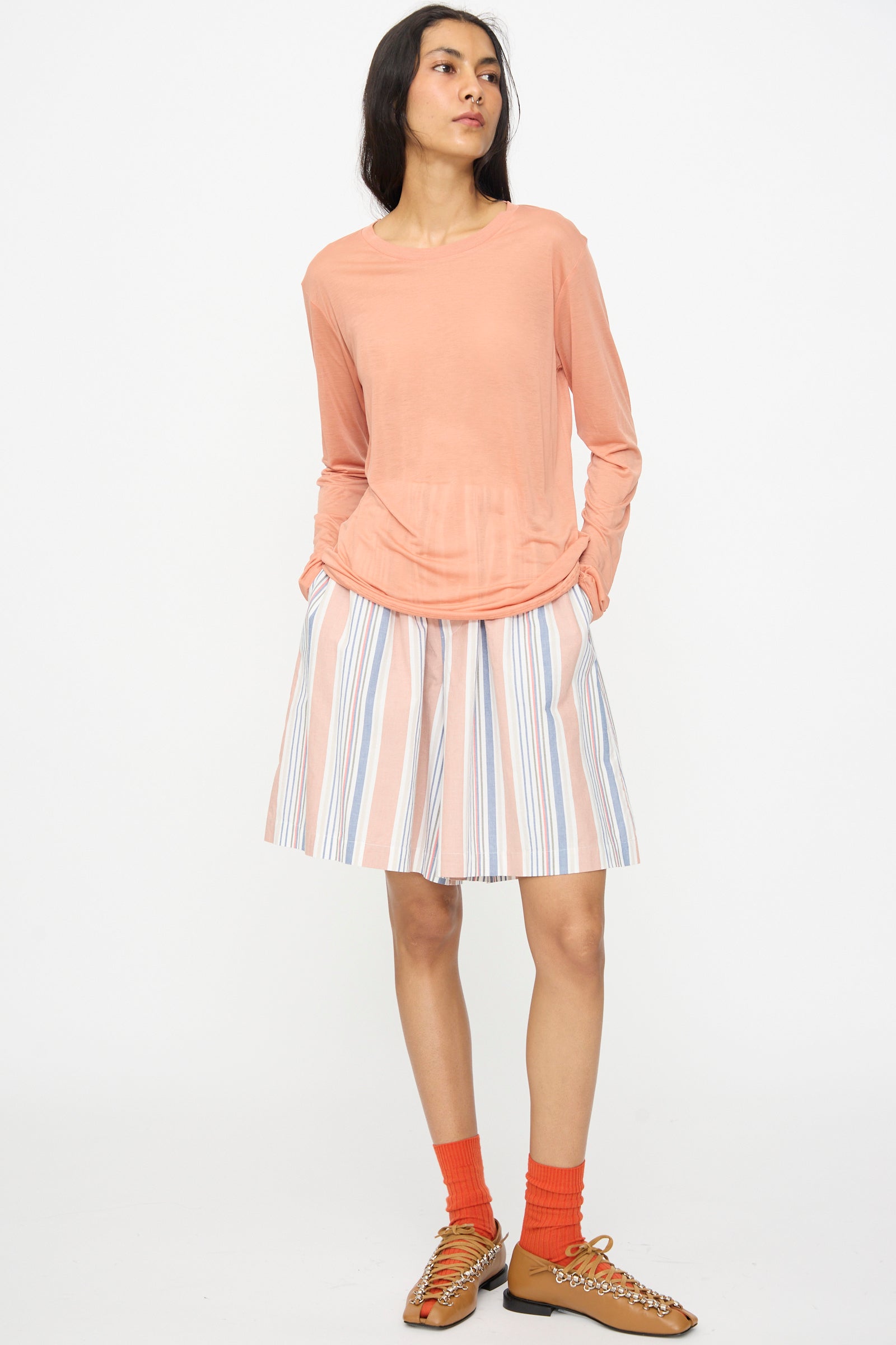 A woman stands with hands in pockets, wearing the Baserange Long Sleeve Tee in Branch Terracotta, a striped skirt, orange socks, and tan shoes with chain details against a plain white background.