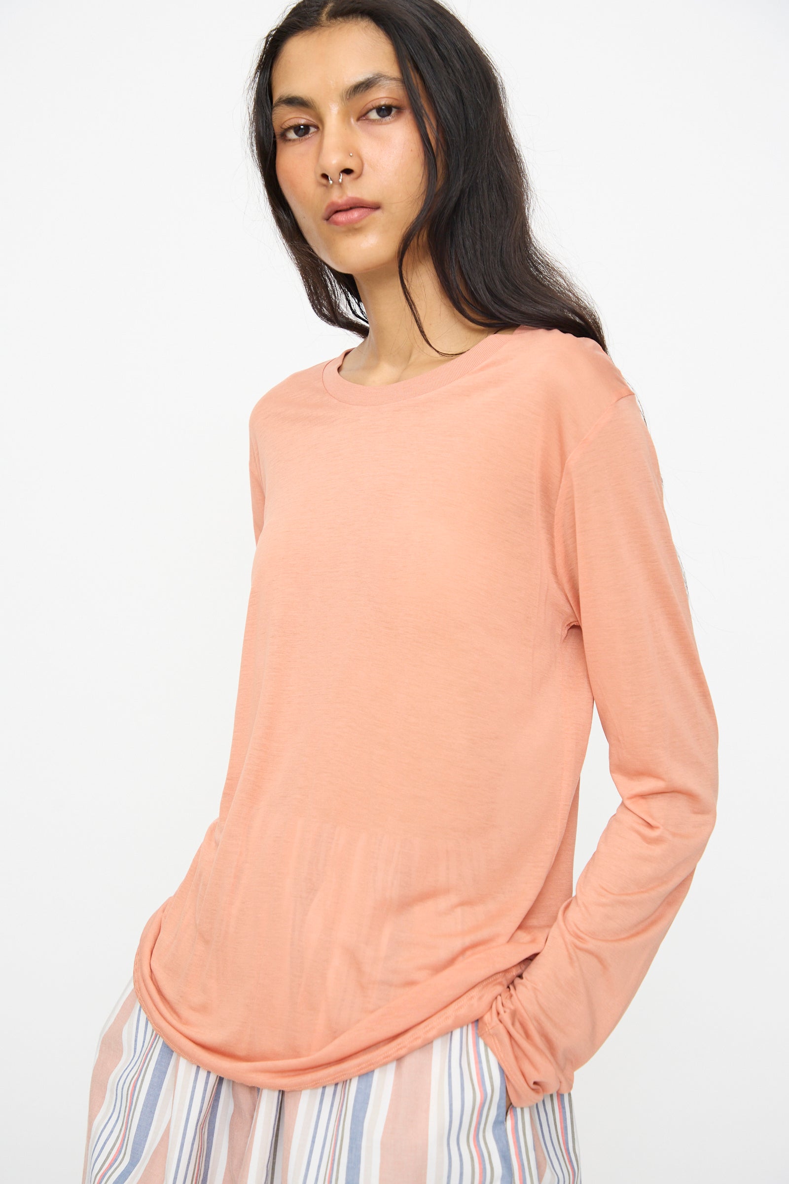 Someone wearing the Baserange Long Sleeve Tee in Branch Terracotta with a pleated skirt featuring vertical pastel stripes stands against a plain white background.