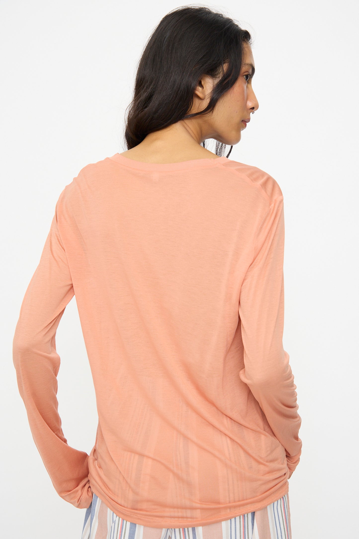 A woman, seen from behind, wears the Baserange Long Sleeve Tee in Branch Terracotta and striped pants, standing against a plain white background.