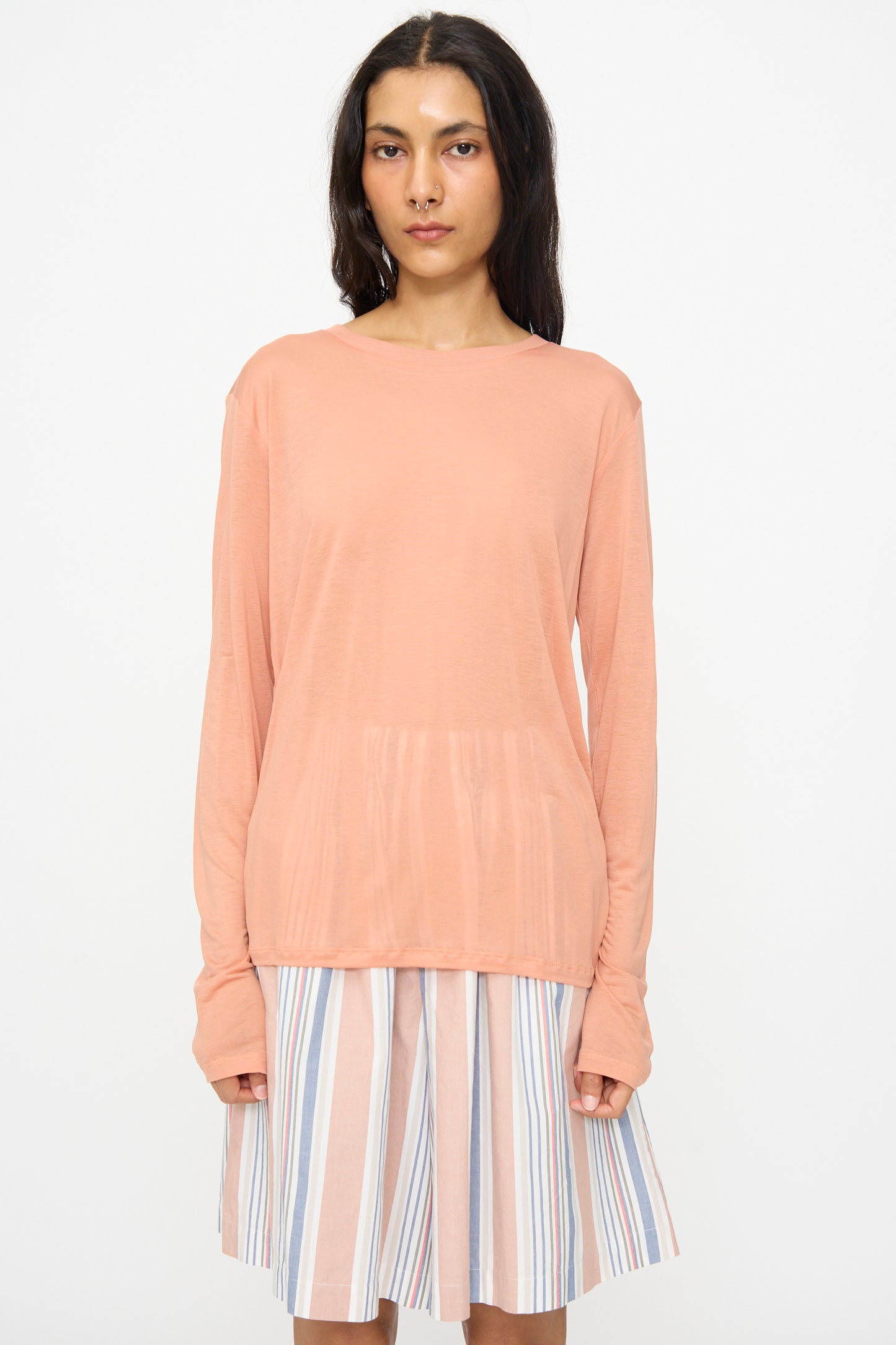 A woman wearing the Baserange Long Sleeve Tee in Branch Terracotta and a pleated, pastel-striped knee-length skirt stands against a plain white background.