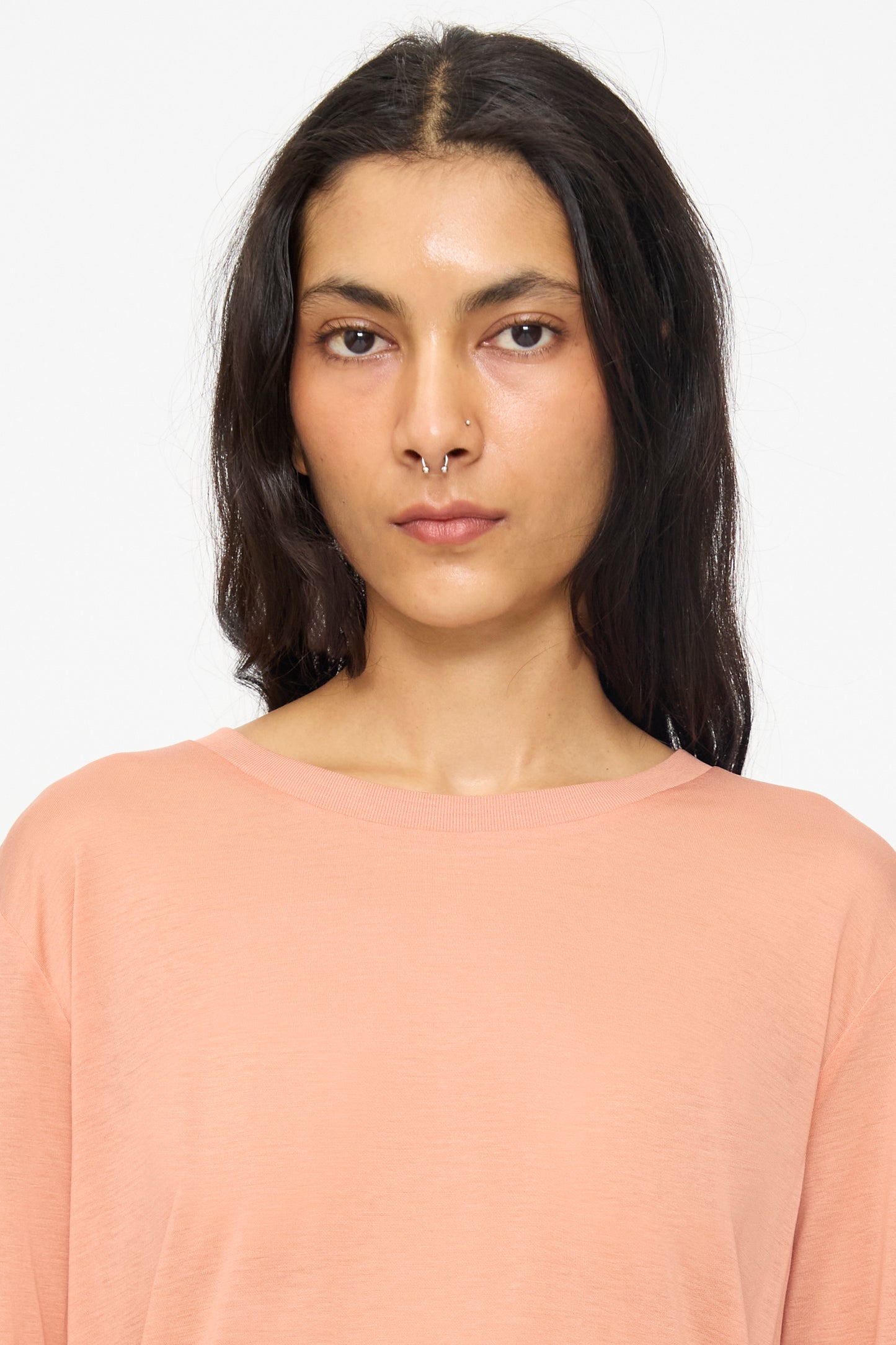A person with long dark hair and a septum piercing wears the Baserange Long Sleeve Tee in Branch Terracotta and looks directly at the camera against a plain background.