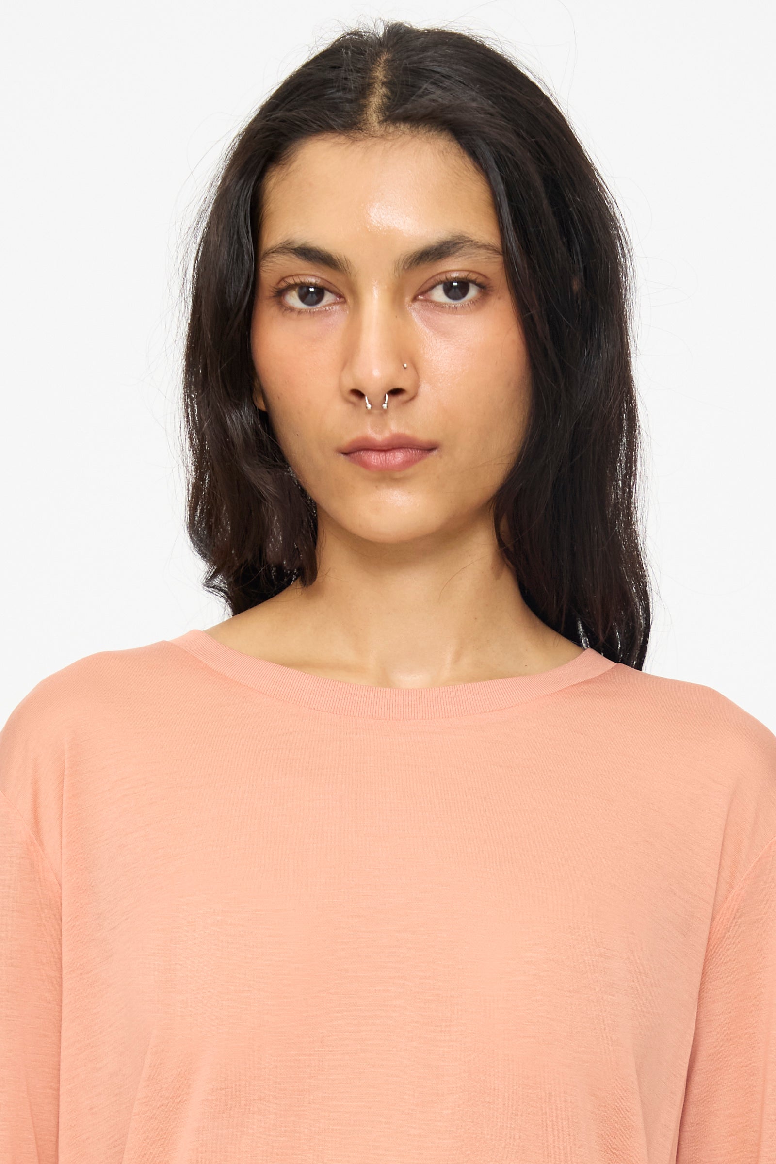 A person with long dark hair and a septum piercing wears the Baserange Long Sleeve Tee in Branch Terracotta and looks directly at the camera against a plain background.