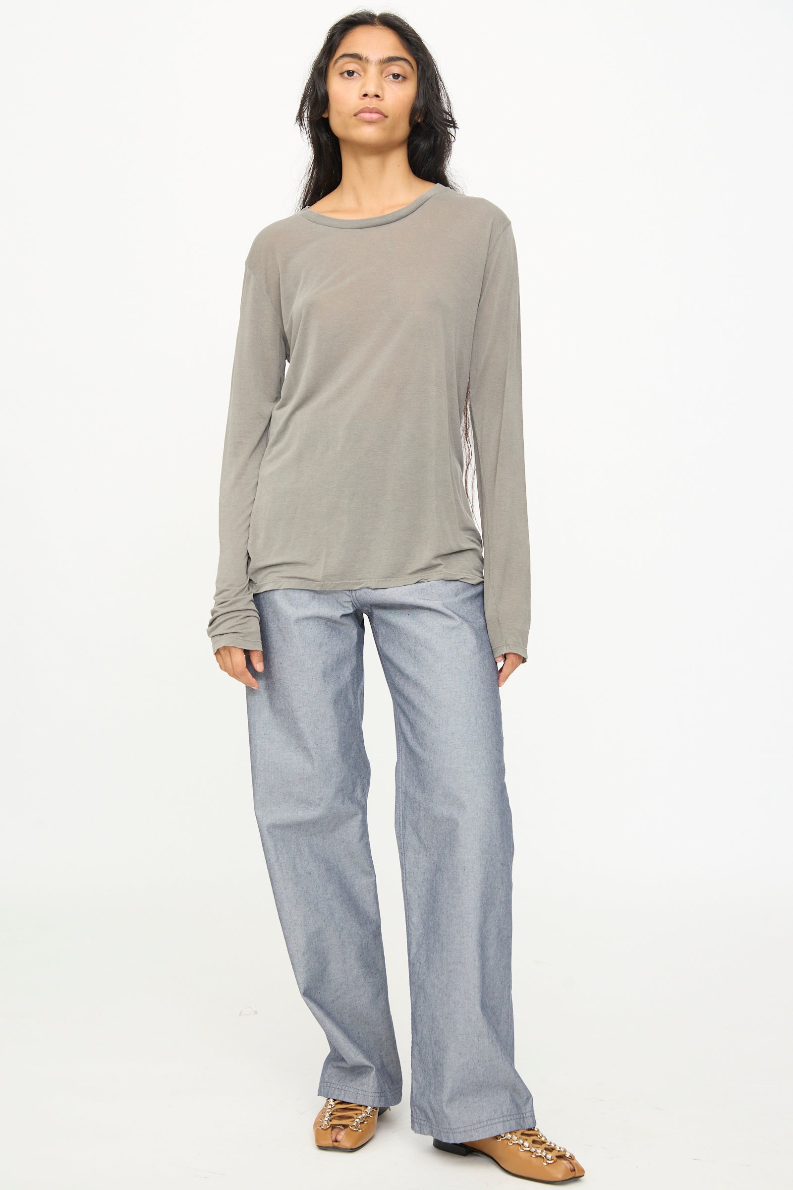 A person stands facing forward wearing the Baserange Long Sleeve Tee in Gilda Grey, paired with loose light blue jeans and brown flats with chain details, against a plain white background.