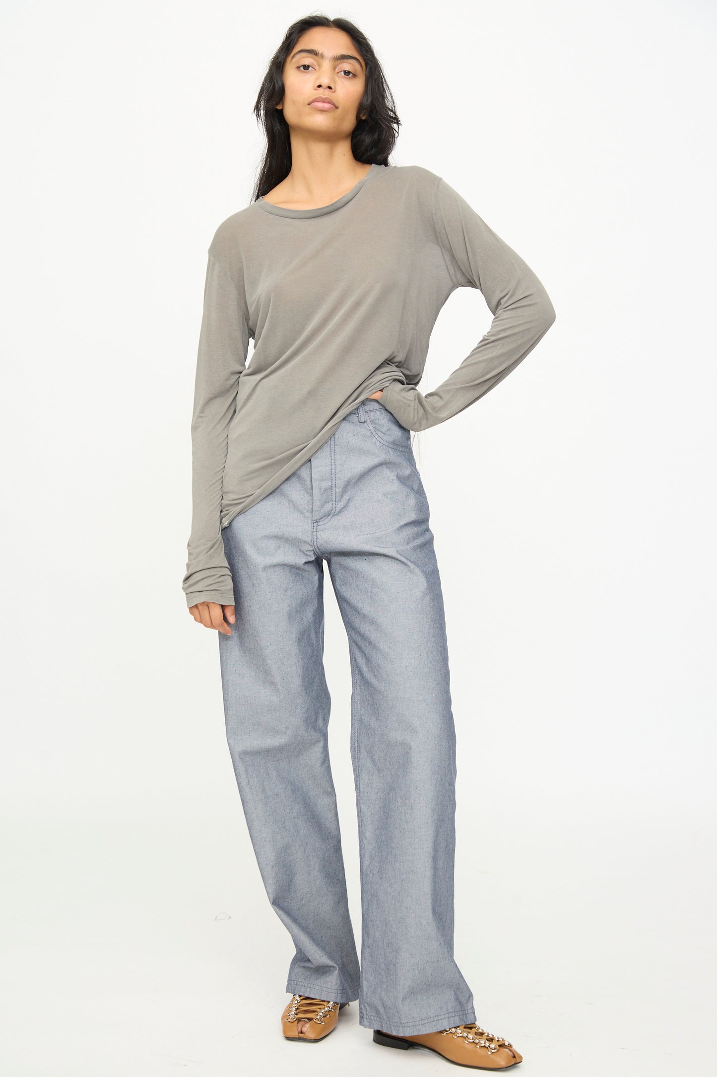 A person stands against a plain background, wearing the Baserange Long Sleeve Tee in Gilda Grey—a loose bamboo lyocell crewneck shirt—paired with wide-leg light blue jeans and brown sandals.