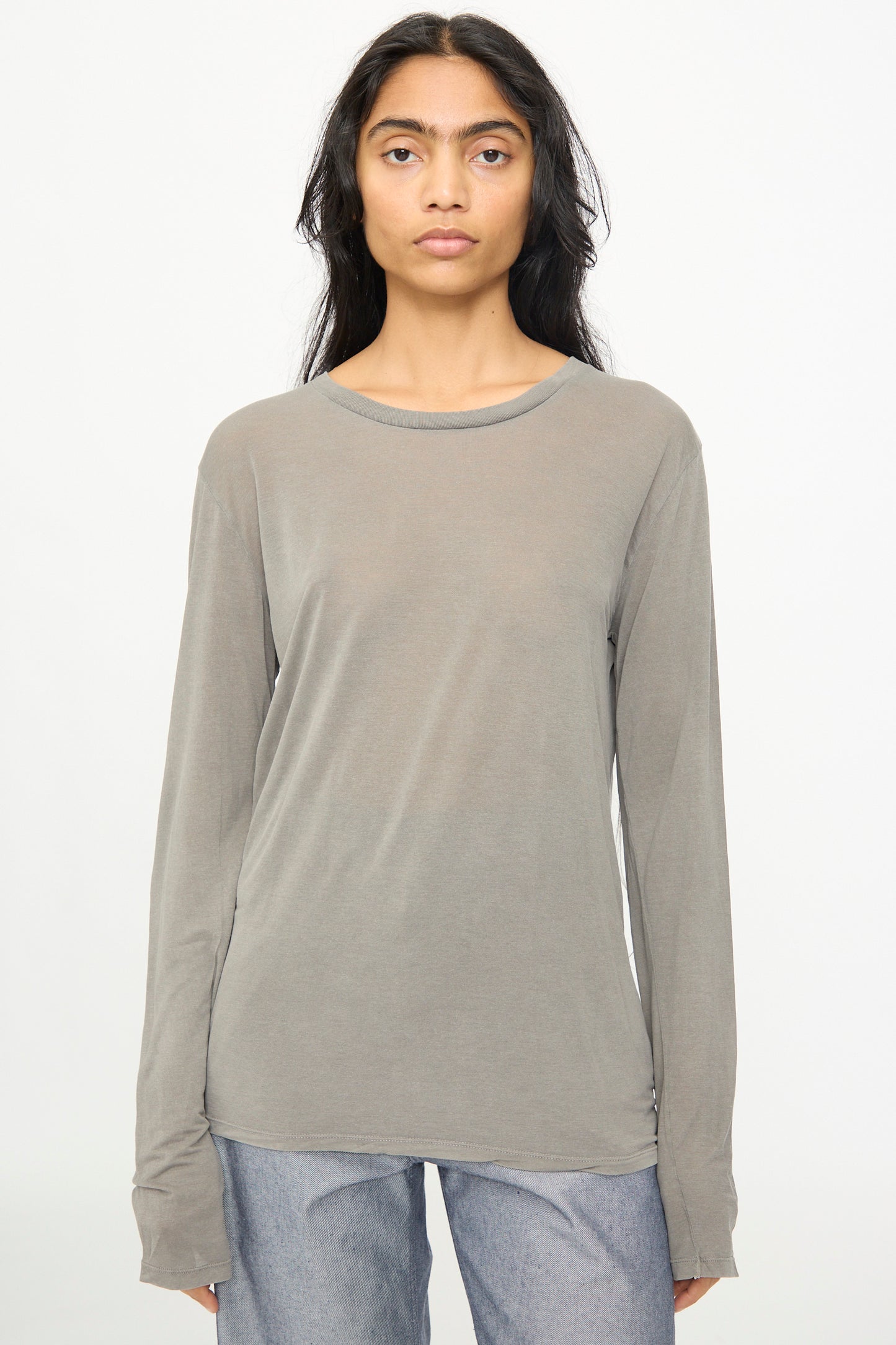 A person with long dark hair wears the Baserange Long Sleeve Tee in Gilda Grey paired with light blue jeans, standing against a plain white background.