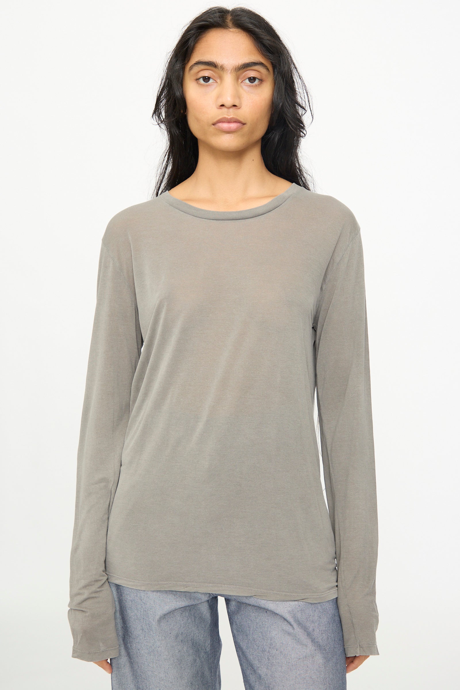 A person with long dark hair wears the Baserange Long Sleeve Tee in Gilda Grey paired with light blue jeans, standing against a plain white background.