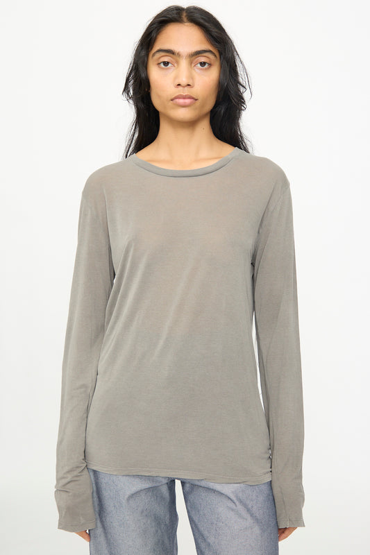 A person with long dark hair wears the Baserange Long Sleeve Tee in Gilda Grey paired with light blue jeans, standing against a plain white background.