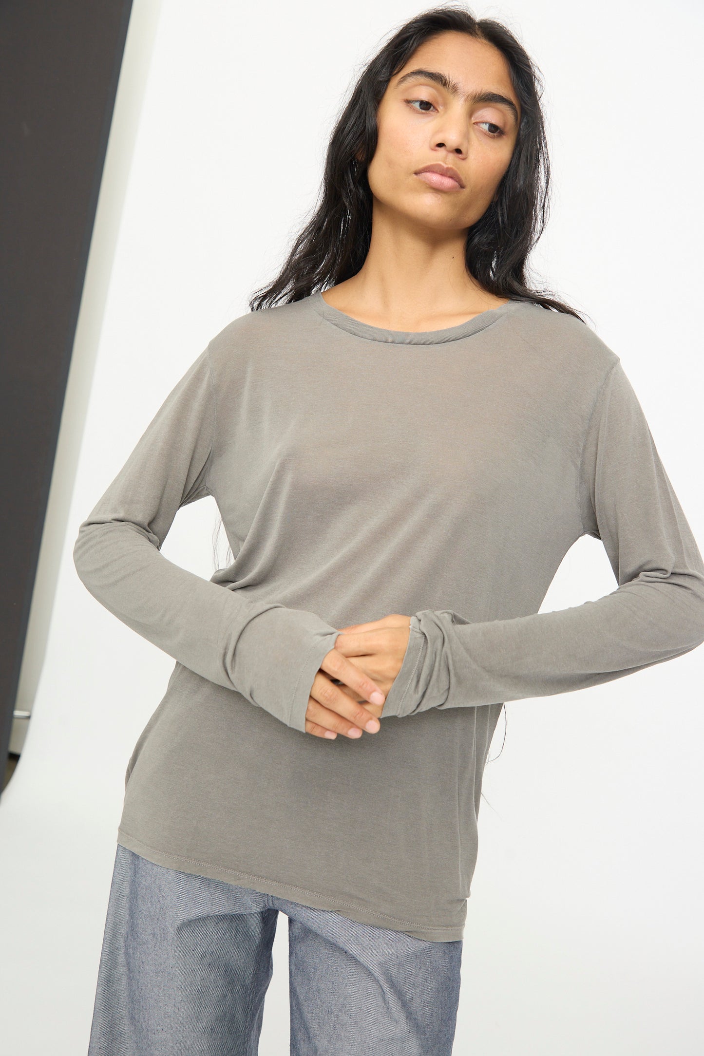 Person with long dark hair wearing the Baserange Long Sleeve Tee in Gilda Grey and gray pants stands against a plain background with hands clasped at waist.