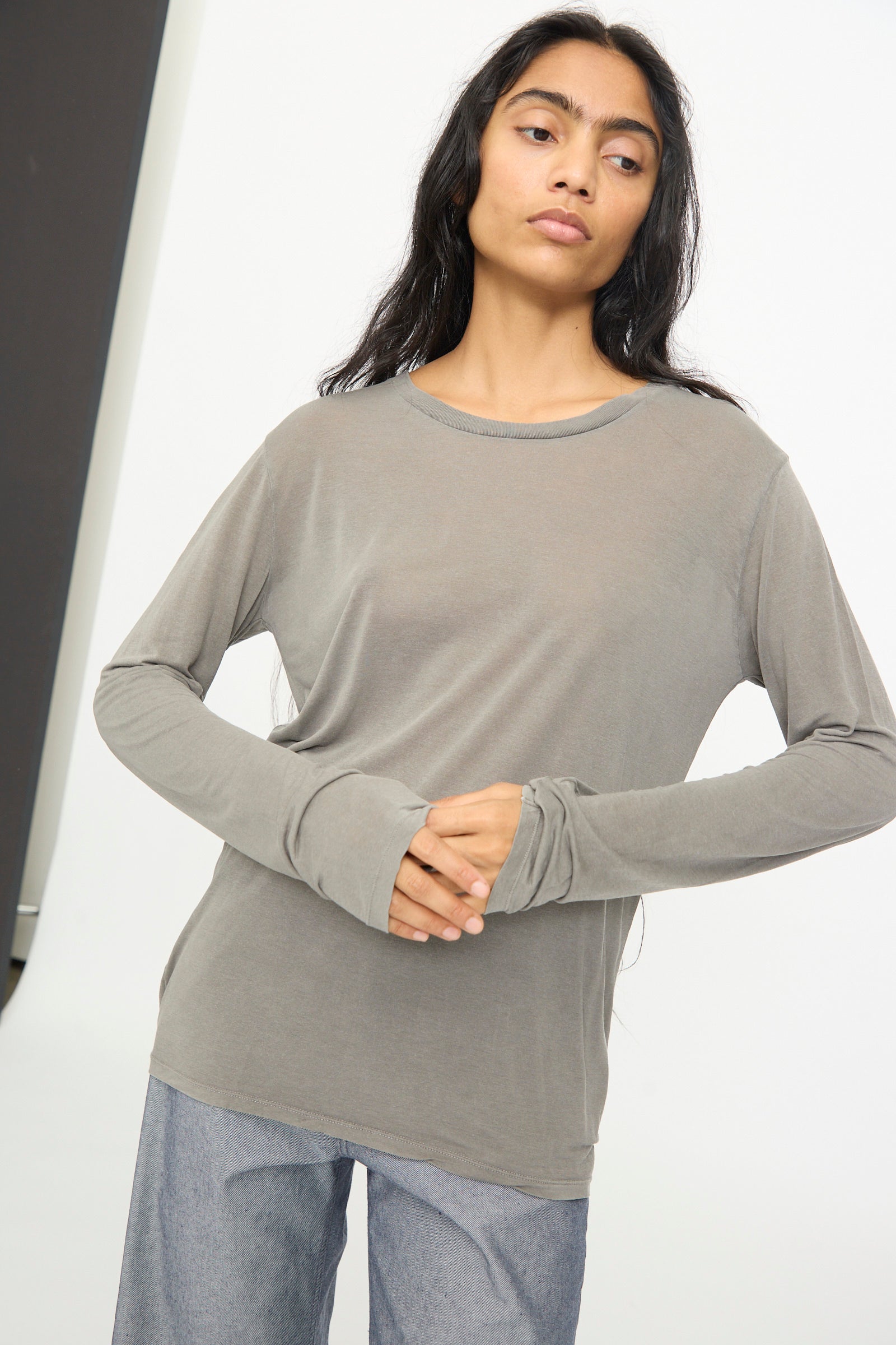 Person with long dark hair wearing the Baserange Long Sleeve Tee in Gilda Grey and gray pants stands against a plain background with hands clasped at waist.