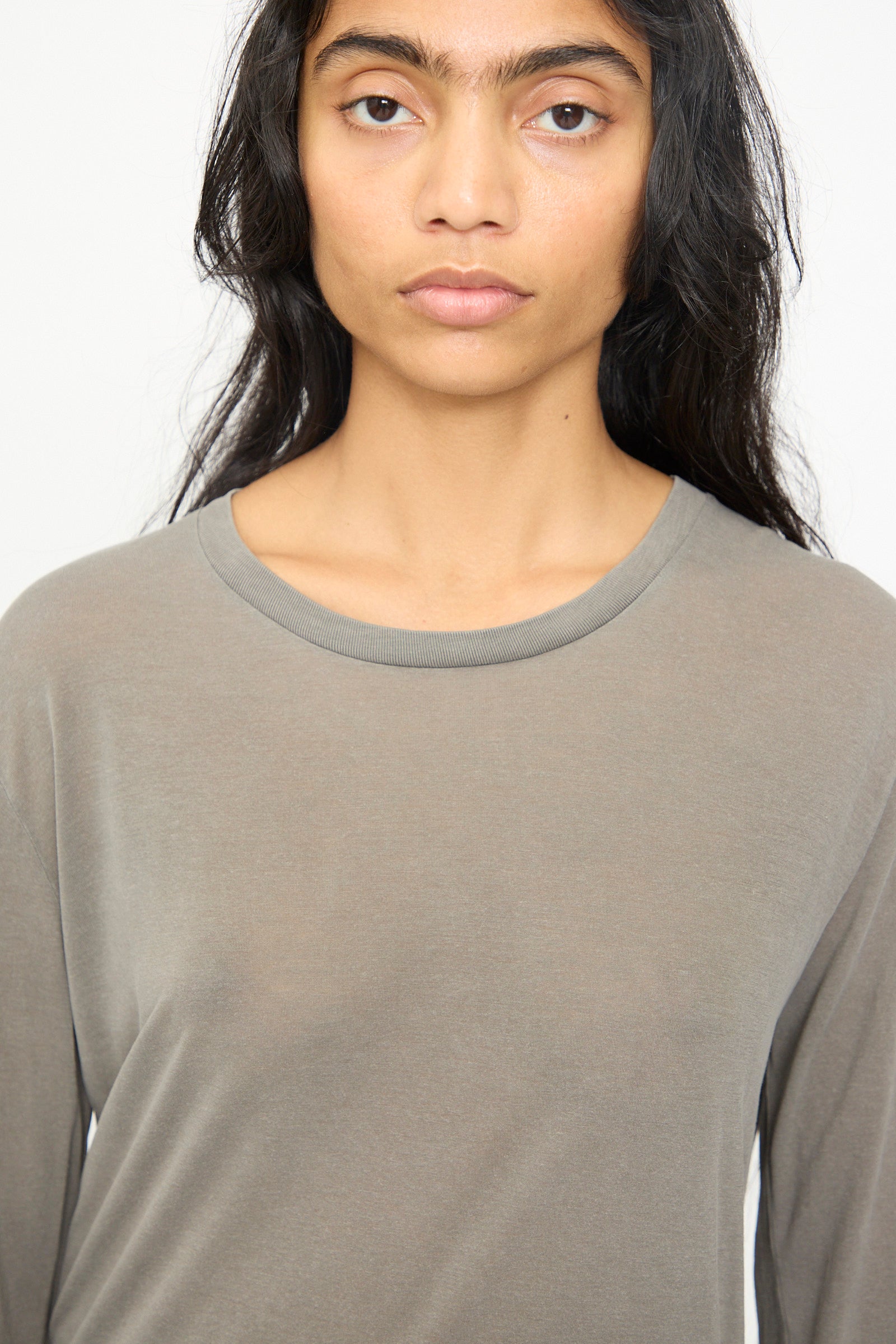 A person with long dark hair is wearing the Baserange Long Sleeve Tee in Gilda Grey, photographed from mid-chest upward against a light background.