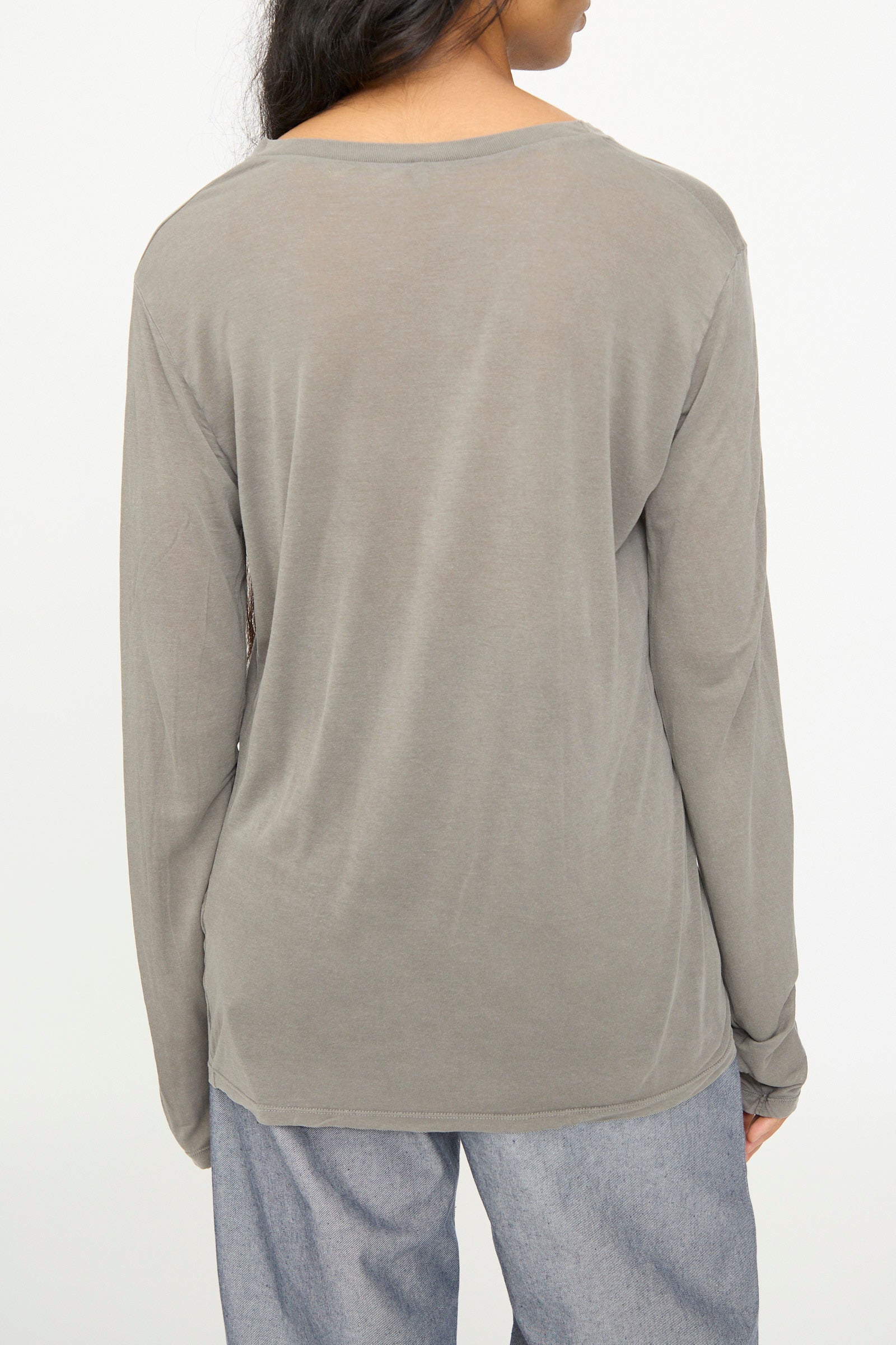 A person wearing the Baserange Long Sleeve Tee in Gilda Grey and light blue pants, photographed from behind against a plain light background.