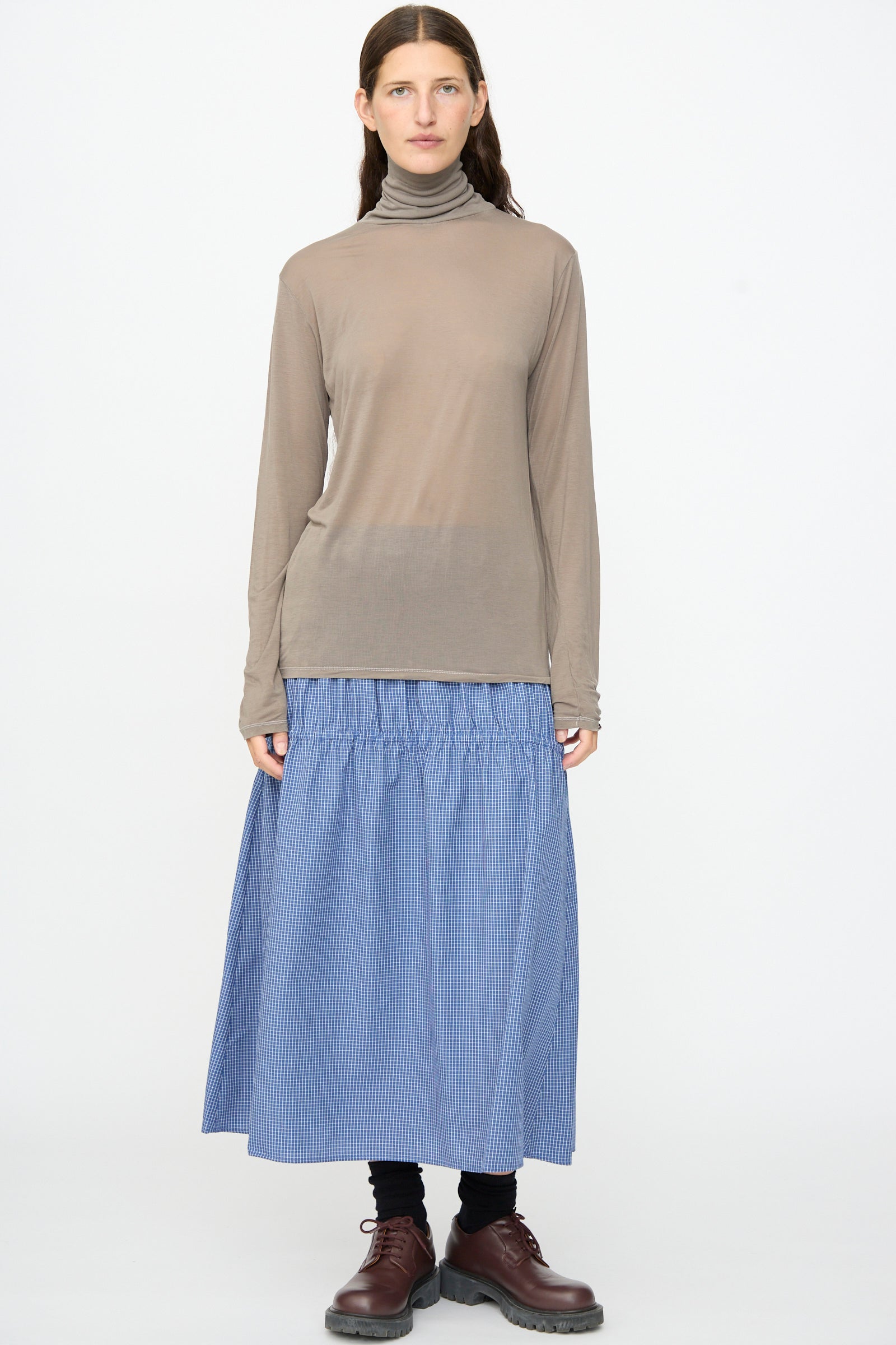 A person wearing the Baserange Long Sleeve Turtleneck in Gilda Grey, a blue gathered midi skirt, black leggings, and brown lace-up shoes, stands against a plain white background.