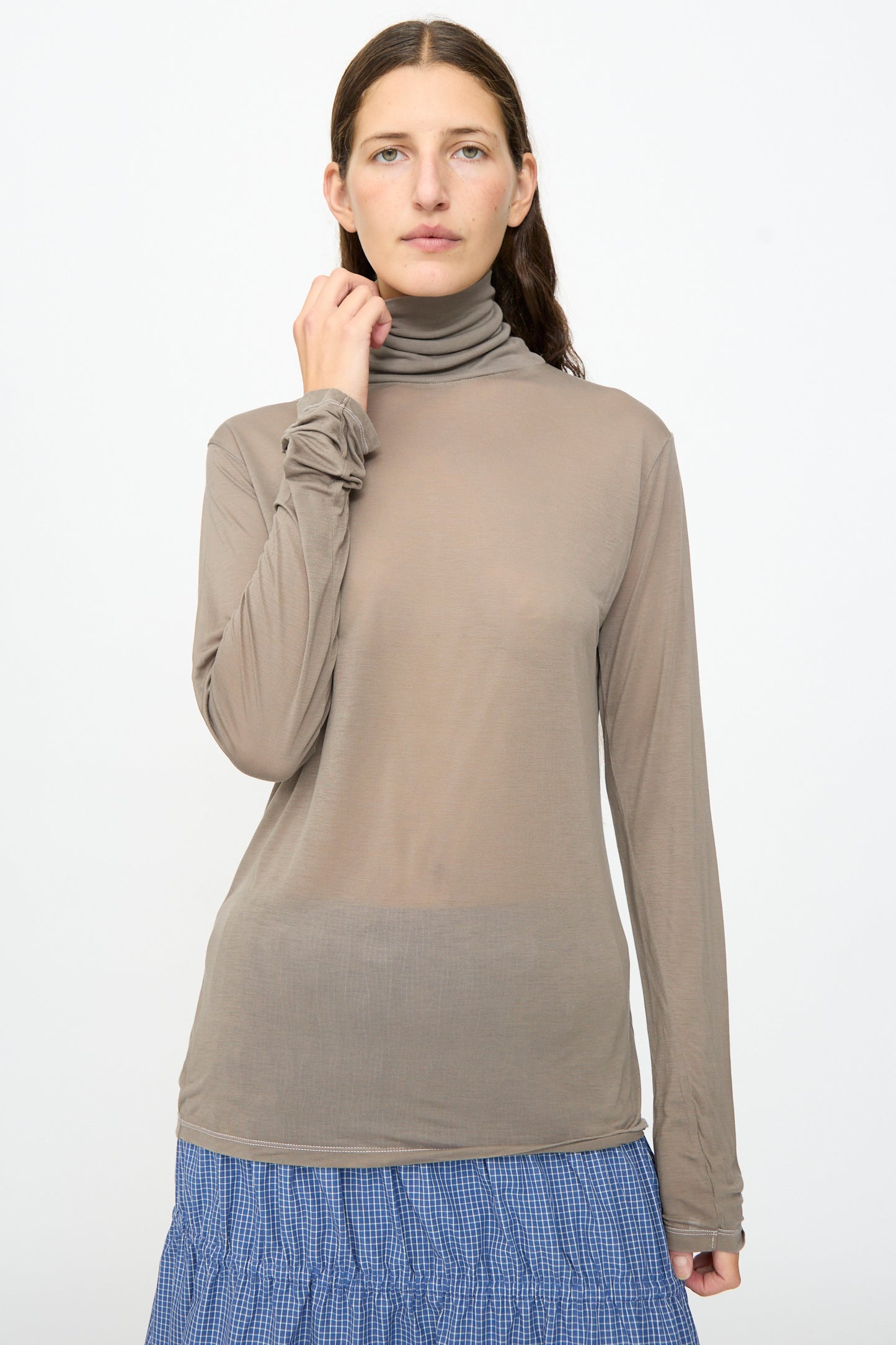 A woman stands against a plain background, wearing the Baserange Long Sleeve Turtleneck in Gilda Grey and a blue checkered skirt, looking directly at the camera with a neutral expression.