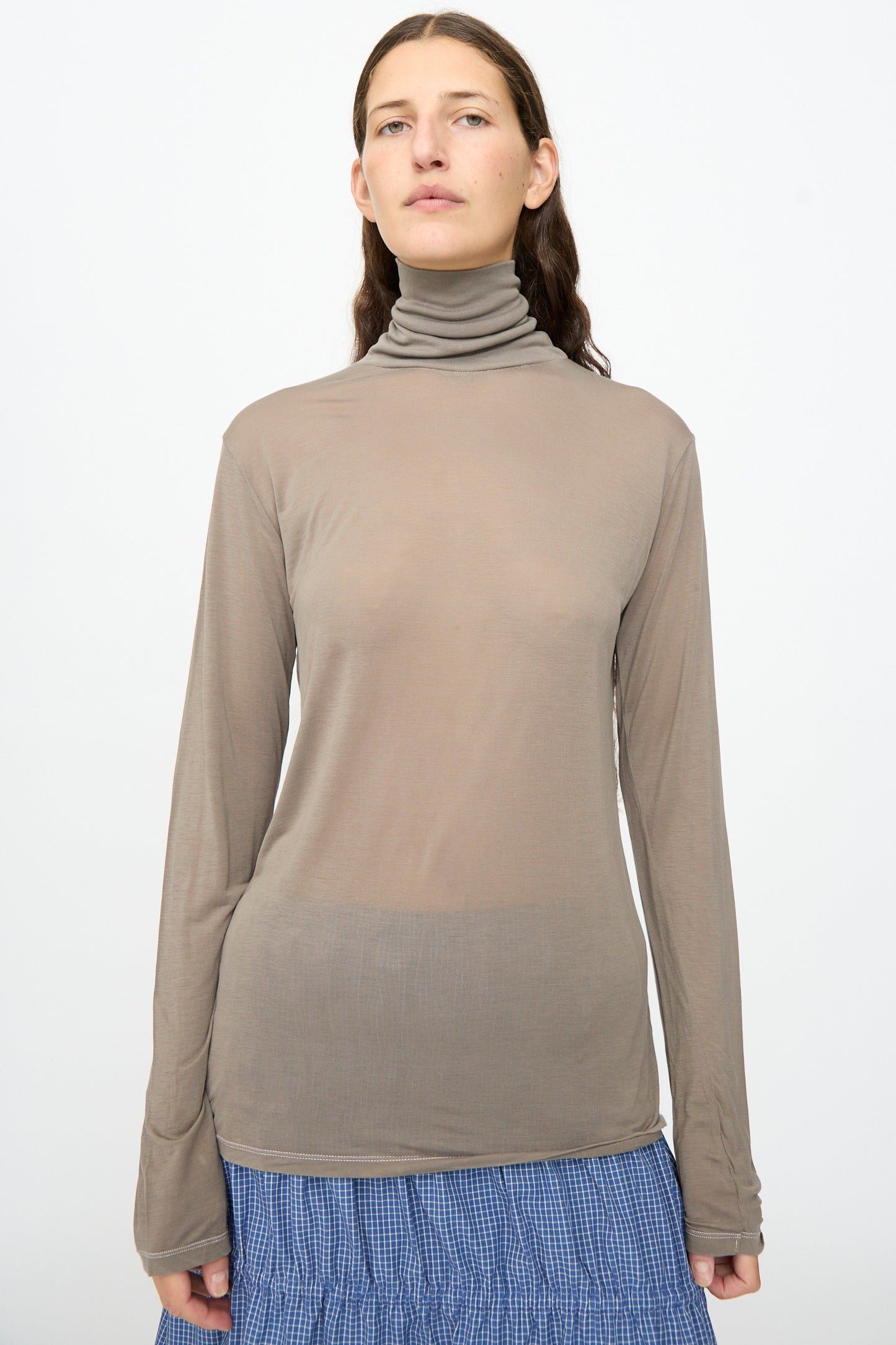 A person with long brown hair wears the Baserange Long Sleeve Turtleneck in Gilda Grey, styled with a blue textured skirt, standing against a plain white background.