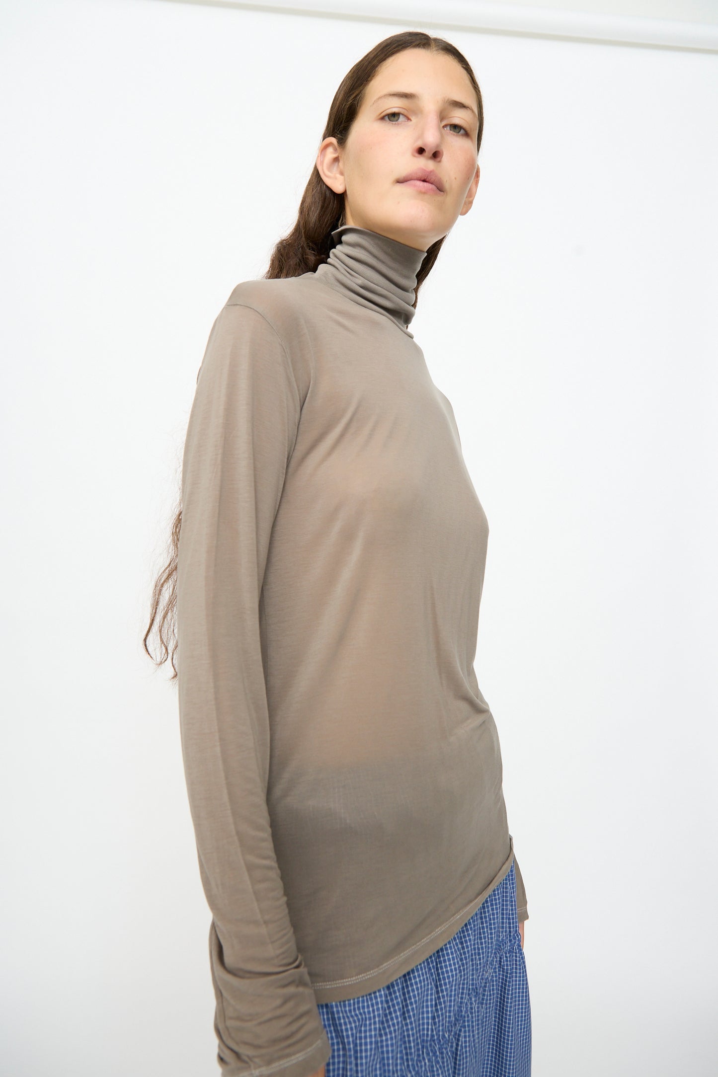 A person with long brown hair wears the Baserange Long Sleeve Turtleneck in Gilda Grey, paired with blue checked pants, standing against a plain white background.