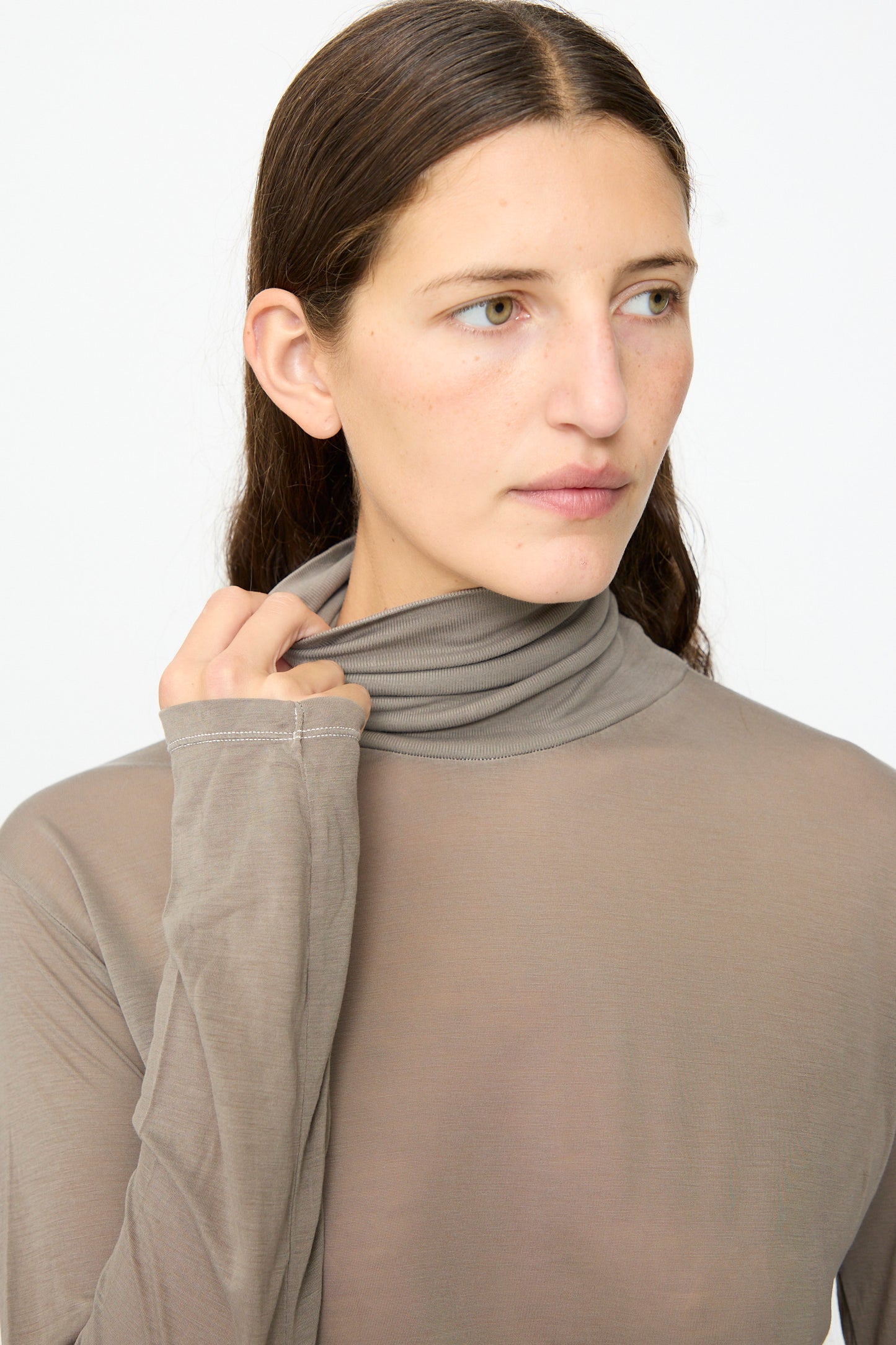A person with long brown hair in the Baserange Long Sleeve Turtleneck in Gilda Grey, styled in a slim fit, looks to the right and holds the collar with one hand against a plain white background.