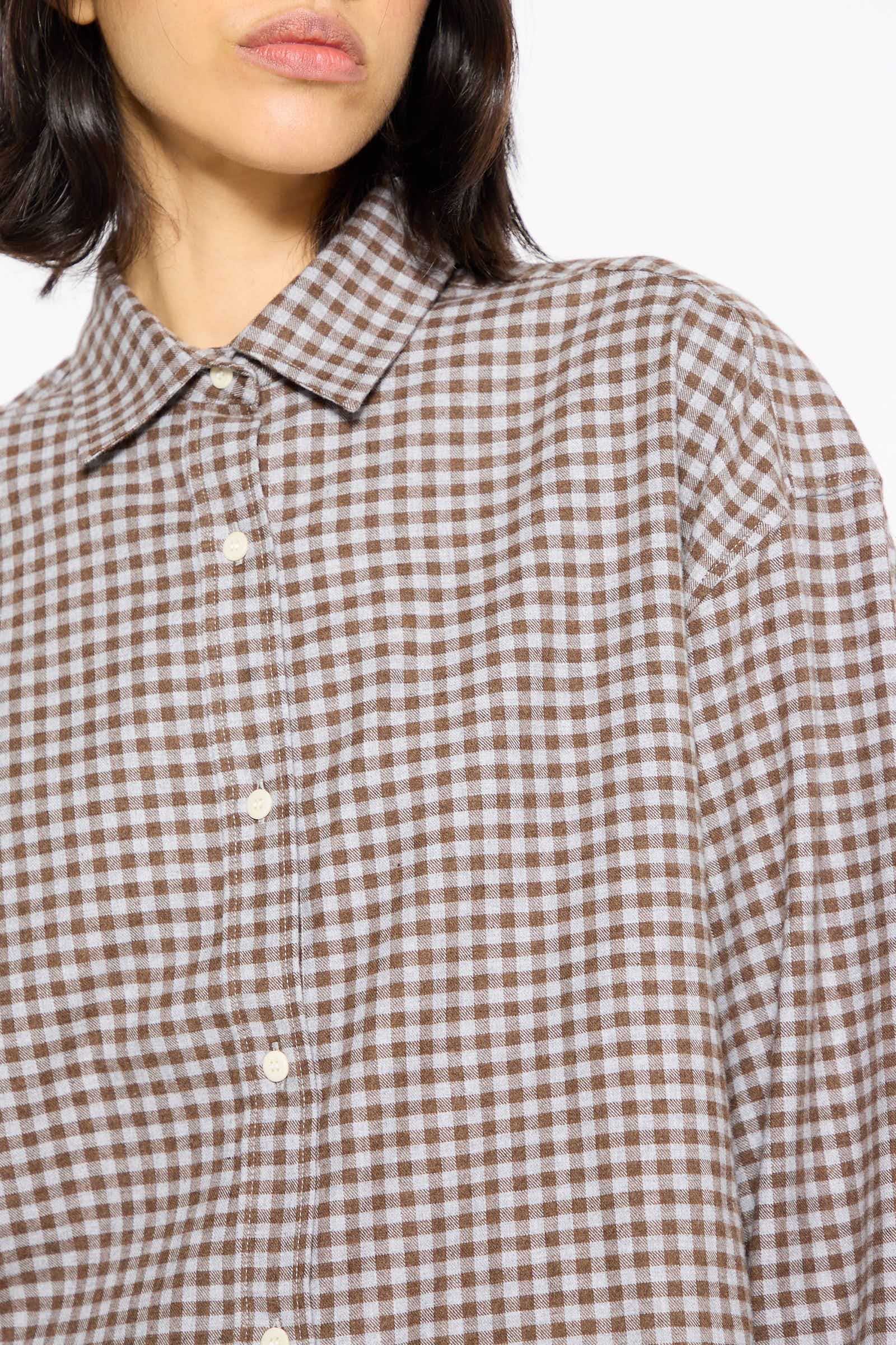 A person wearing the Loose Ole Shirt in Brown Grey Flannel by Baserange, featuring a brown and white checkered pattern, is shown from the shoulders up.