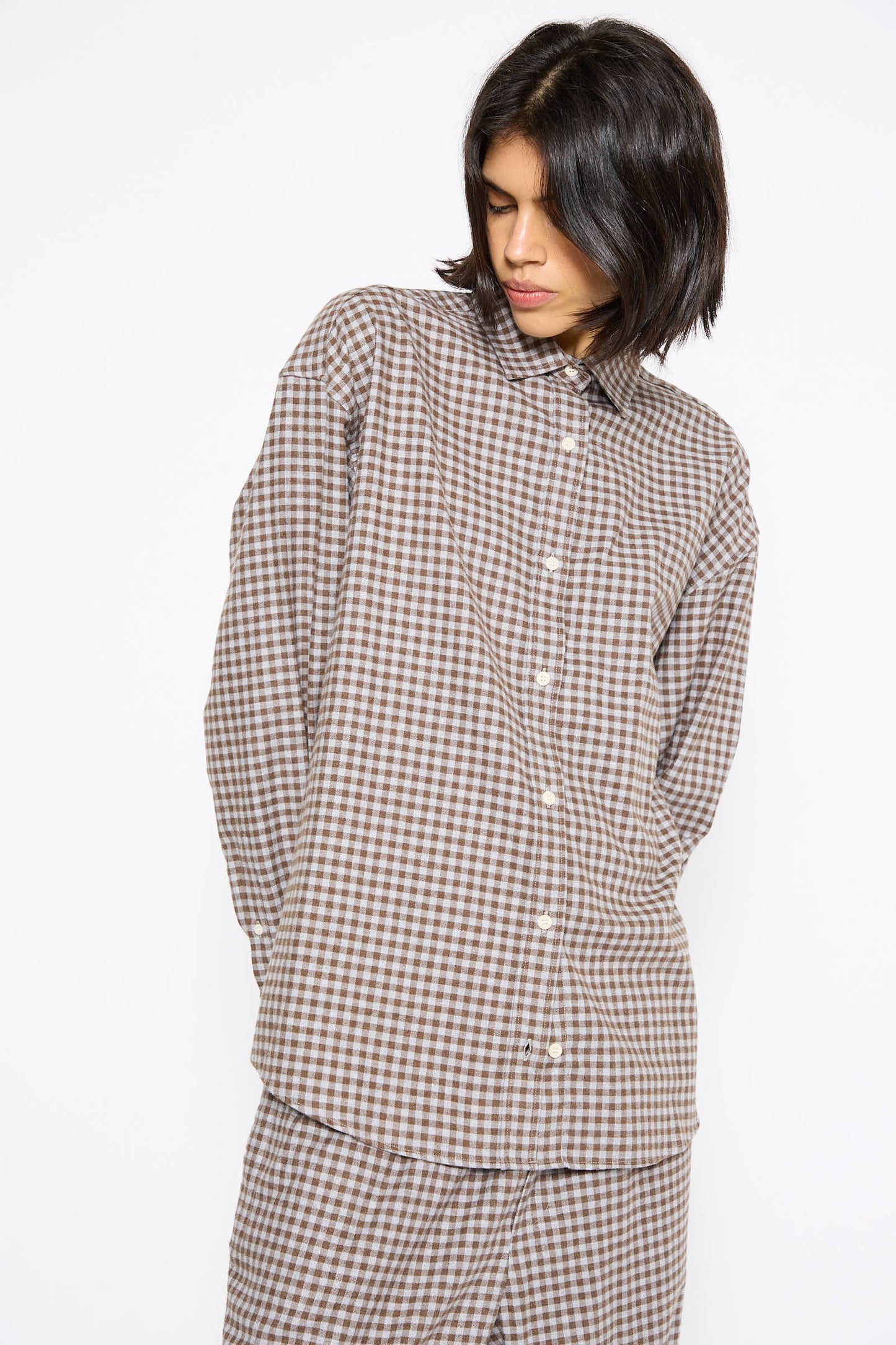 A person with medium-length dark hair is standing against a plain white background, wearing the Loose Ole Shirt in Brown Grey Flannel by Baserange.