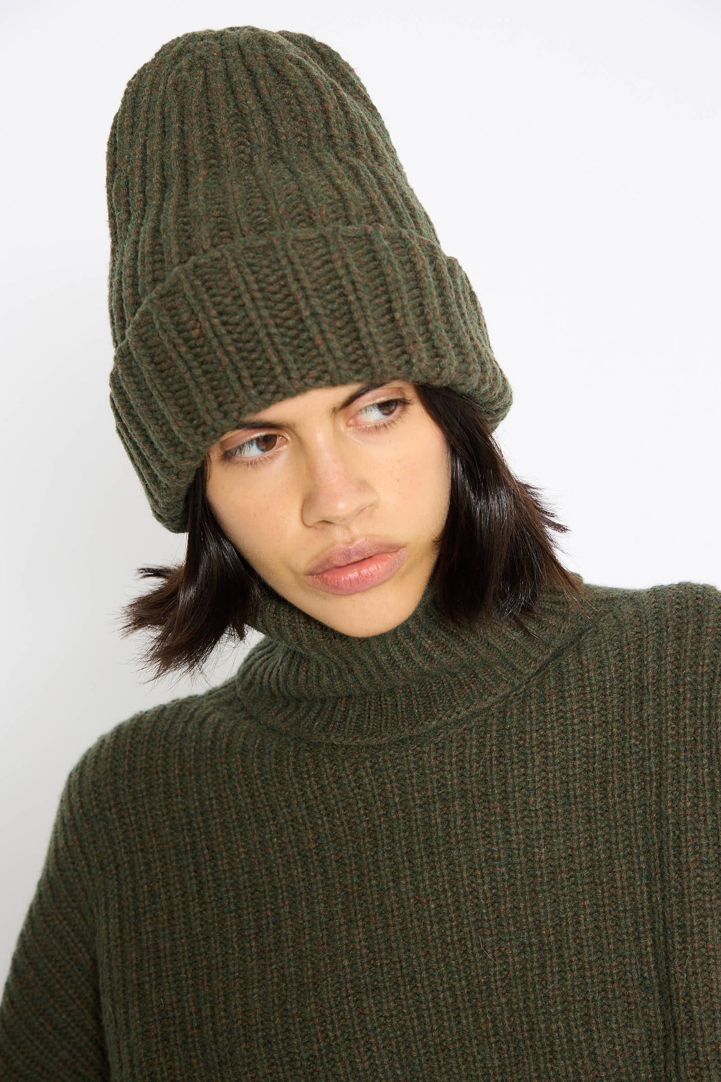 A person wearing a chunky ribbed Mea Beanie in Foret by Baserange and a matching green knit sweater made from recycled wool gazes off to the side.