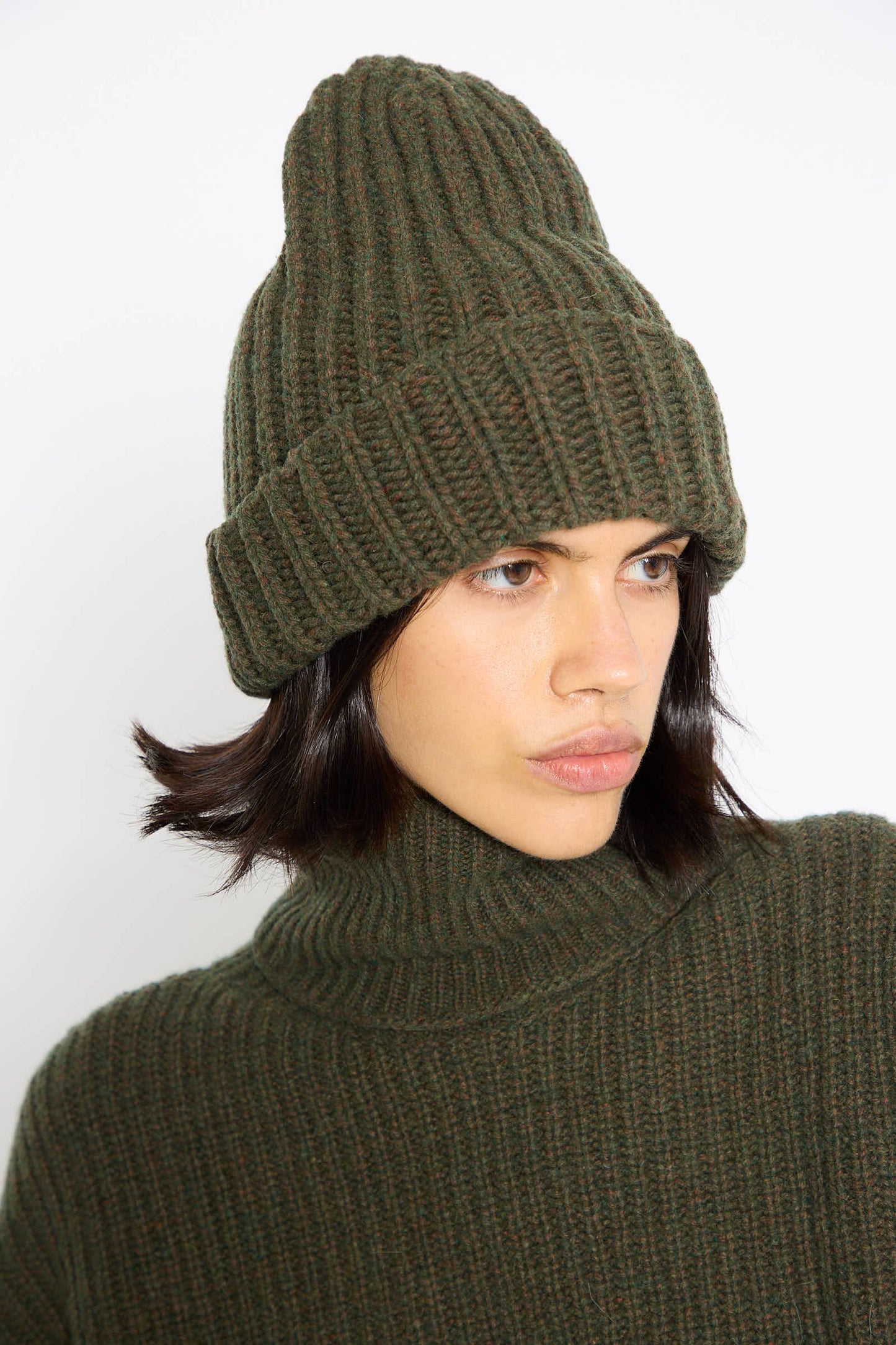 A person wearing the Baserange Mea Beanie in Foret and a matching green ribbed turtleneck sweater looks to the side.