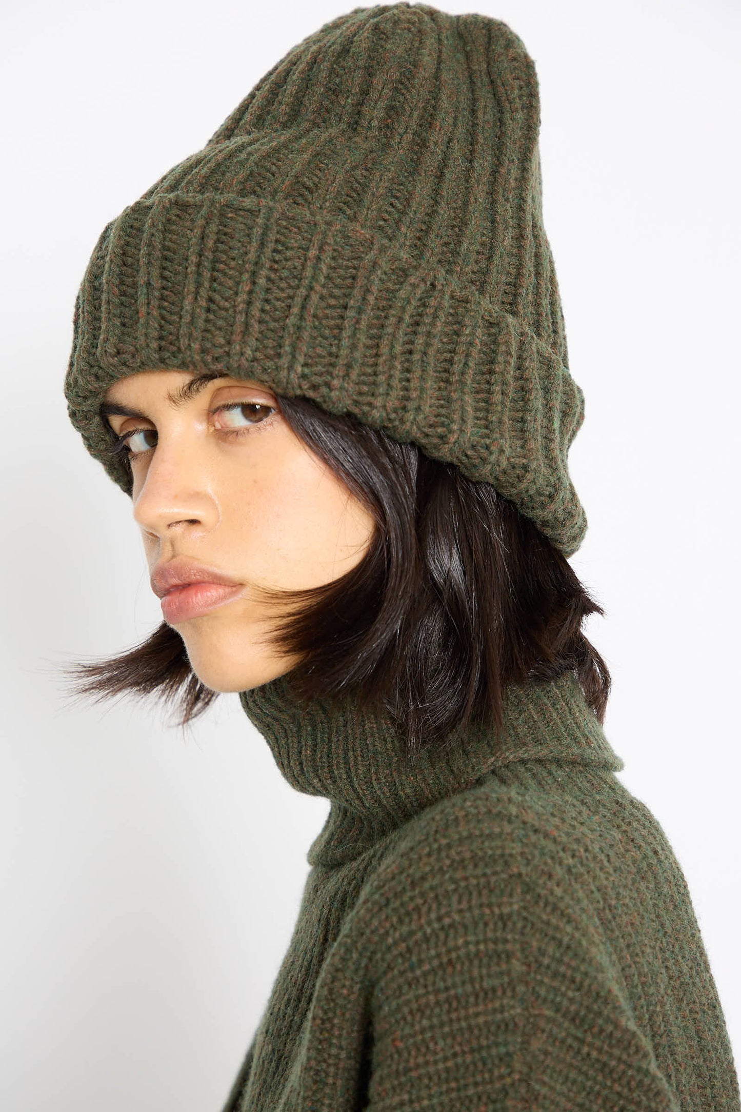 A person wearing an oversized Baserange Mea Beanie in Foret and a matching recycled wool sweater looks sideways with a neutral expression.