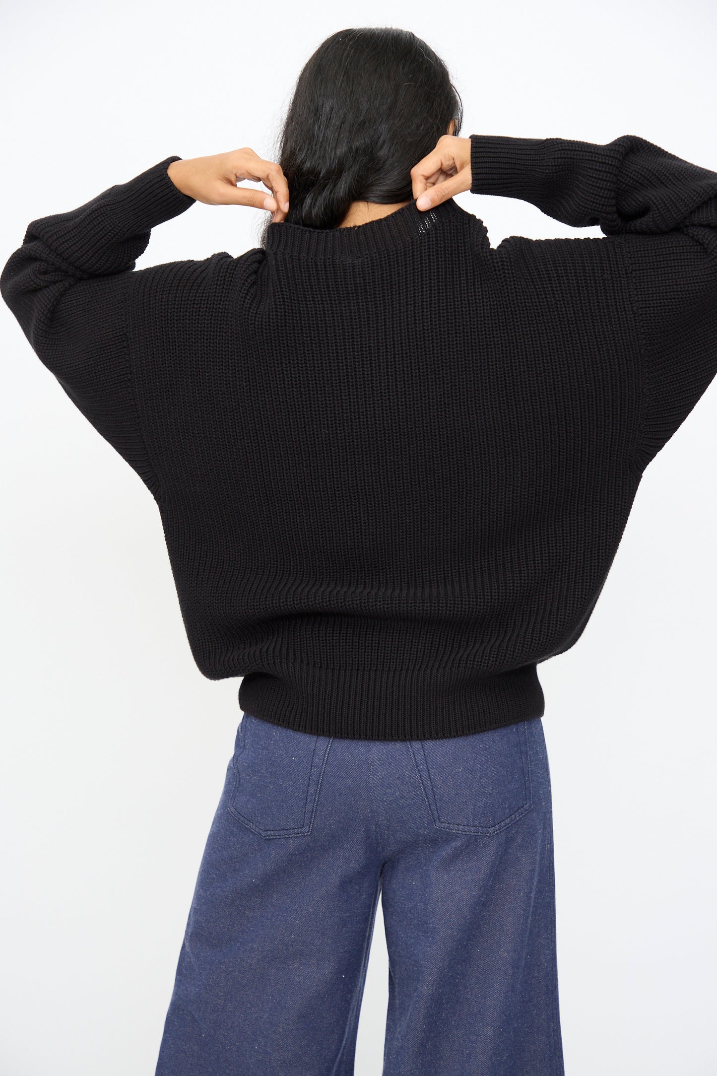 A person stands against a plain white background, adjusting the collar of the Baserange Mea Knit Pullover in Black, an oversized rib-knit sweater, paired with blue wide-leg pants.