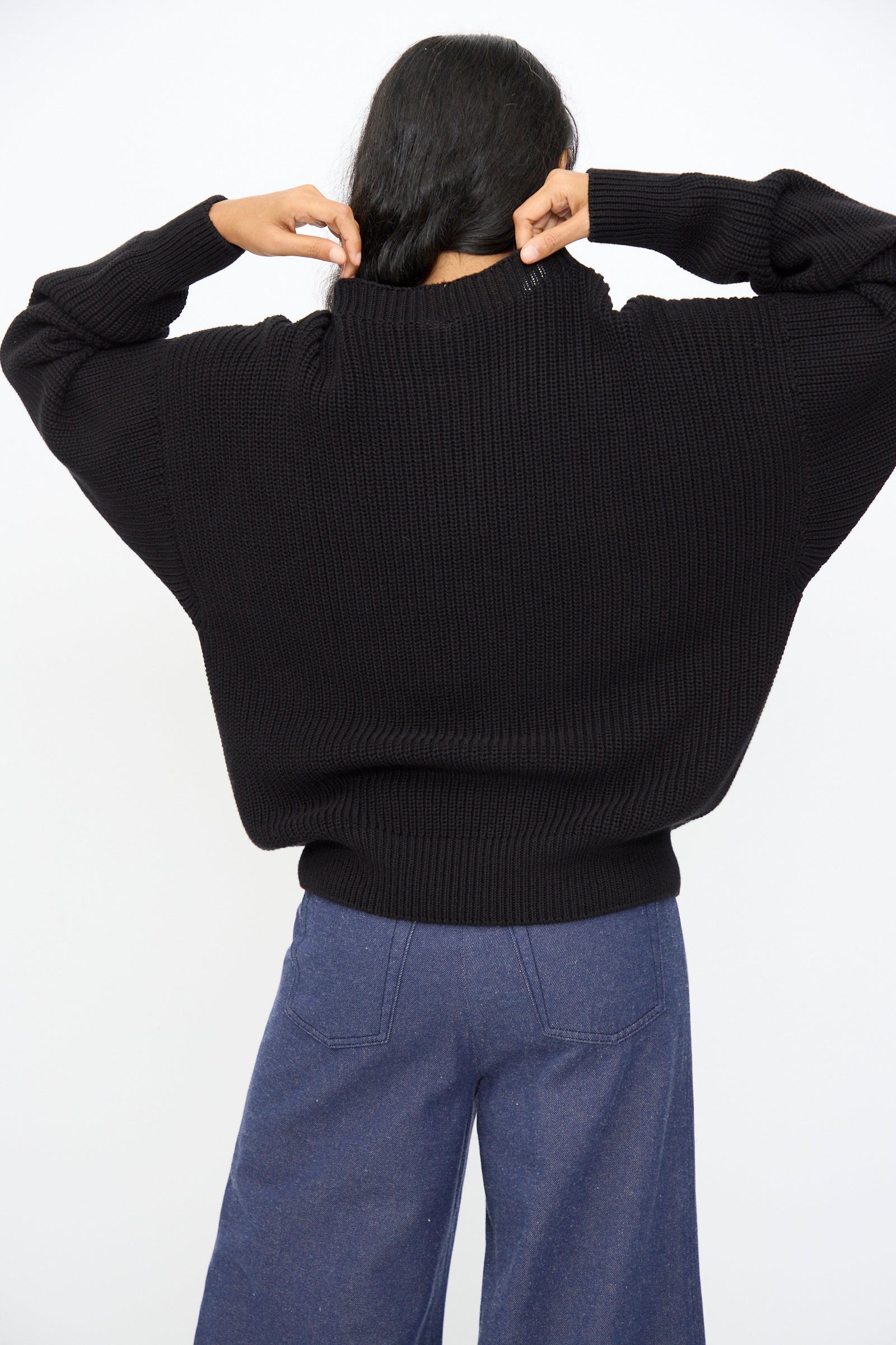 A person stands against a plain white background, adjusting the collar of the Baserange Mea Knit Pullover in Black, an oversized rib-knit sweater, paired with blue wide-leg pants.