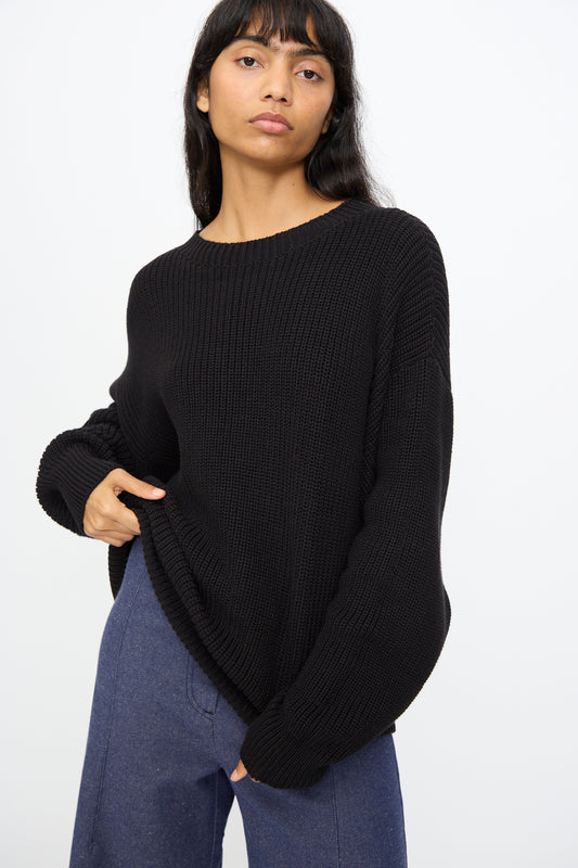 A woman wears the Baserange Mea Knit Pullover in Black with blue pants, standing against a plain white background.