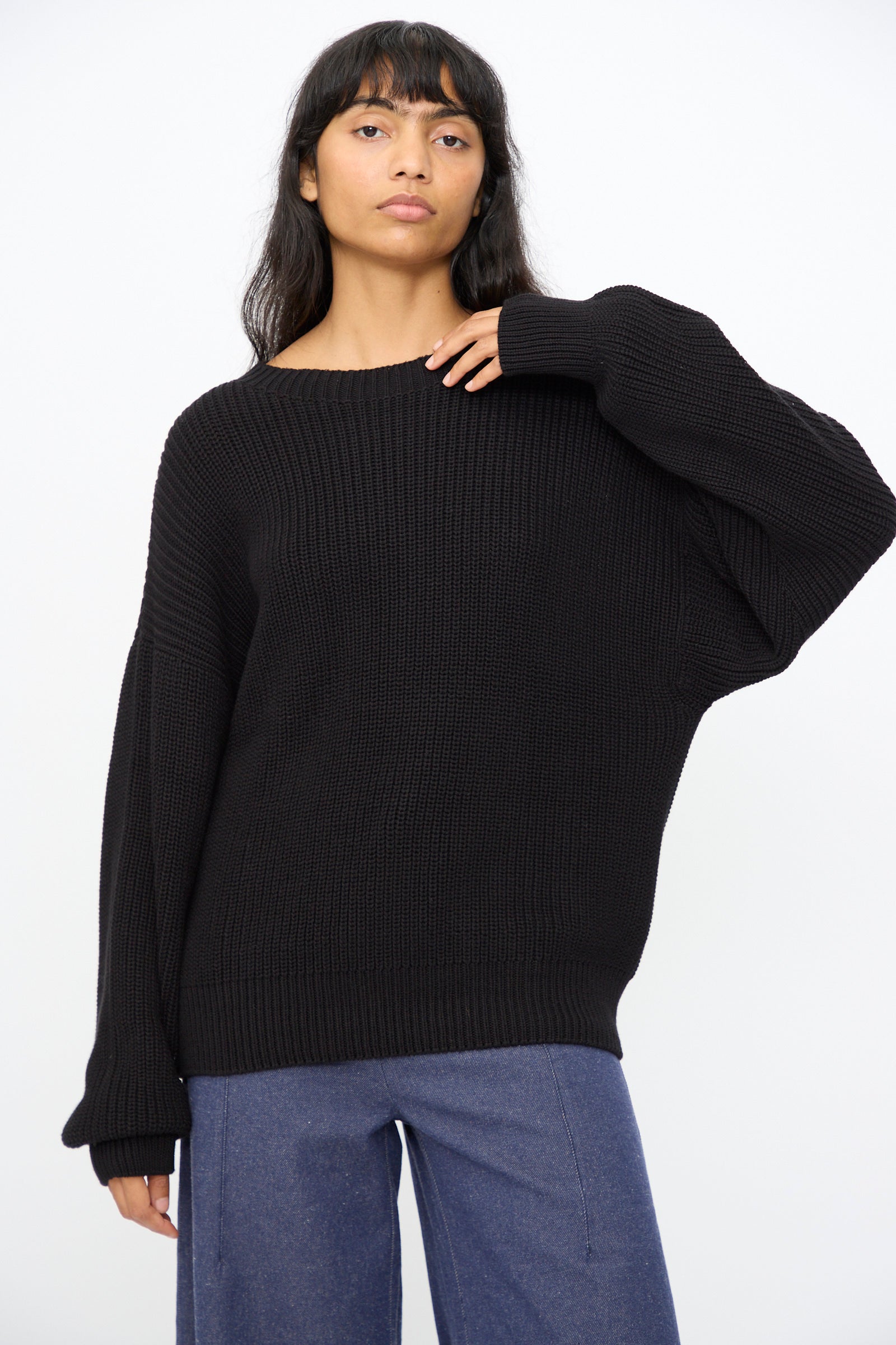 A person wearing the Baserange Mea Knit Pullover in Black with an oversized fit and blue pants stands against a plain white background.