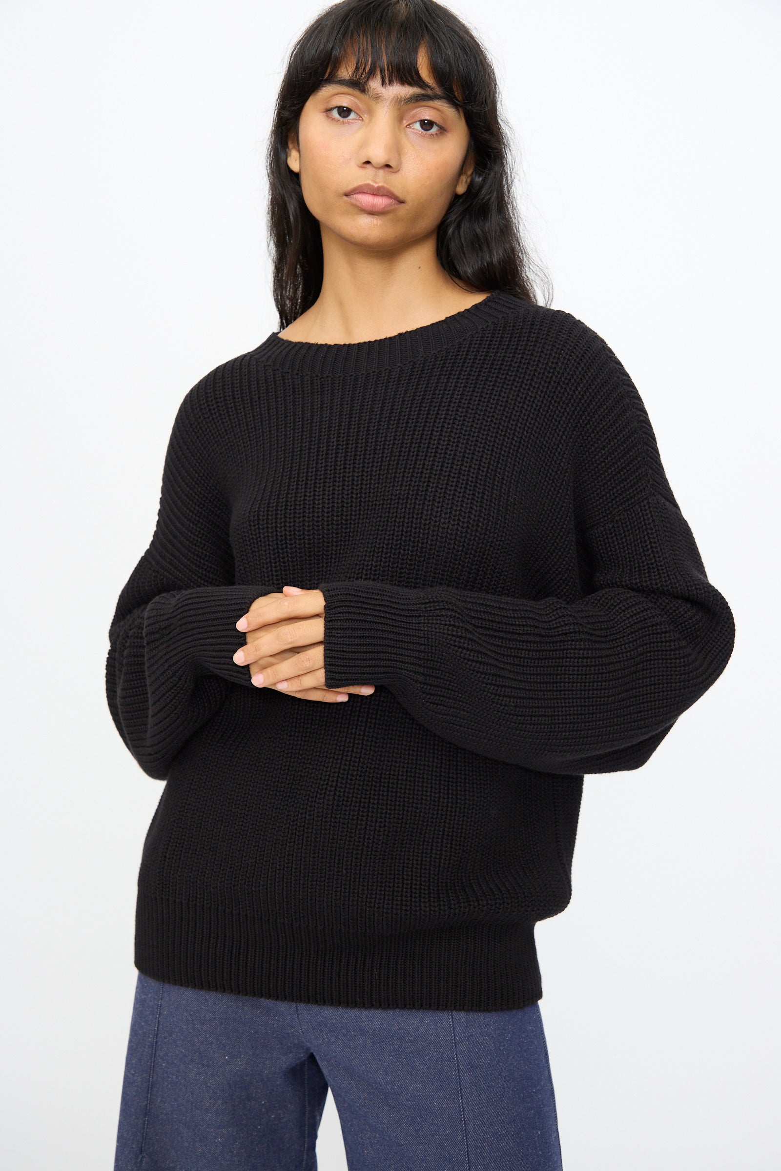 A person with long dark hair wears the Baserange Mea Knit Pullover in Black styled oversized with blue pants, standing against a plain white background.