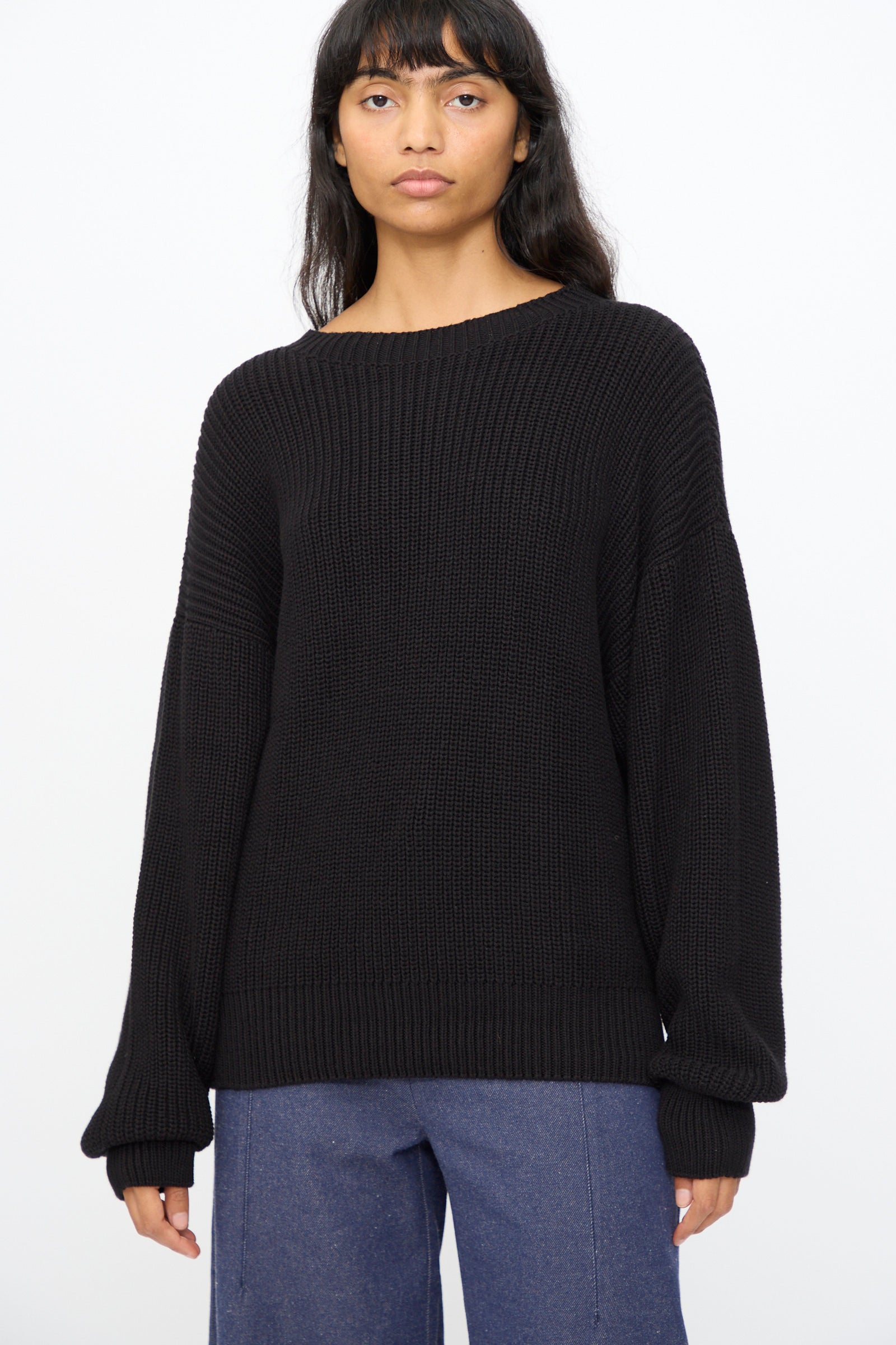 A person with long dark hair wears the Baserange Mea Knit Pullover in Black, styled oversized, paired with blue pants against a plain light background.
