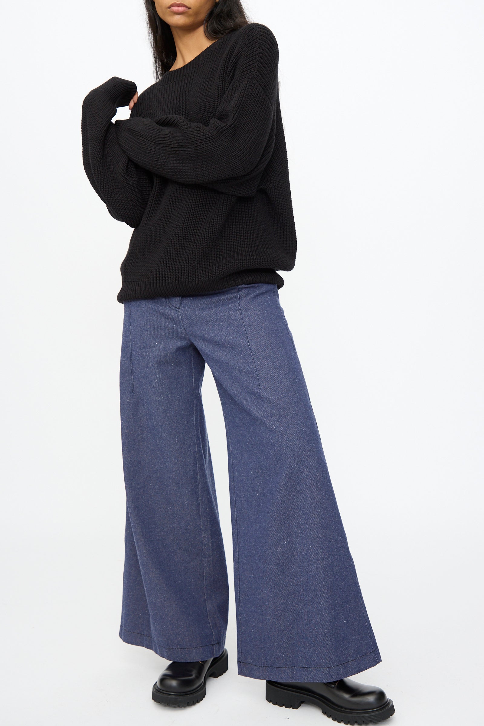 A person wearing the Baserange Mea Knit Pullover in Black, styled with wide-leg blue jeans and black platform shoes, stands against a plain white background.