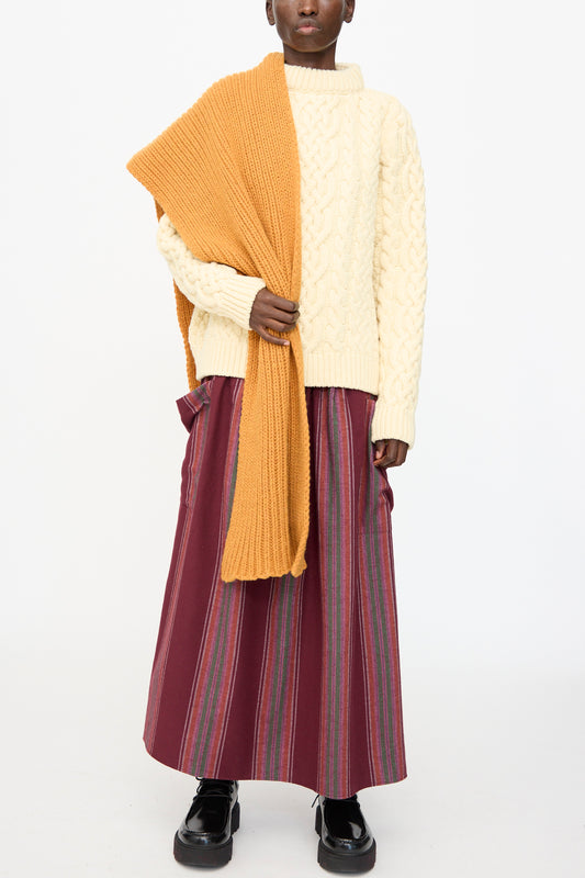 A person wears a cream cable-knit sweater, black shoes, striped maroon maxi skirt, and a Baserange Mea Scarf in Clair Orange—made in France from recycled wool—against a plain white background.