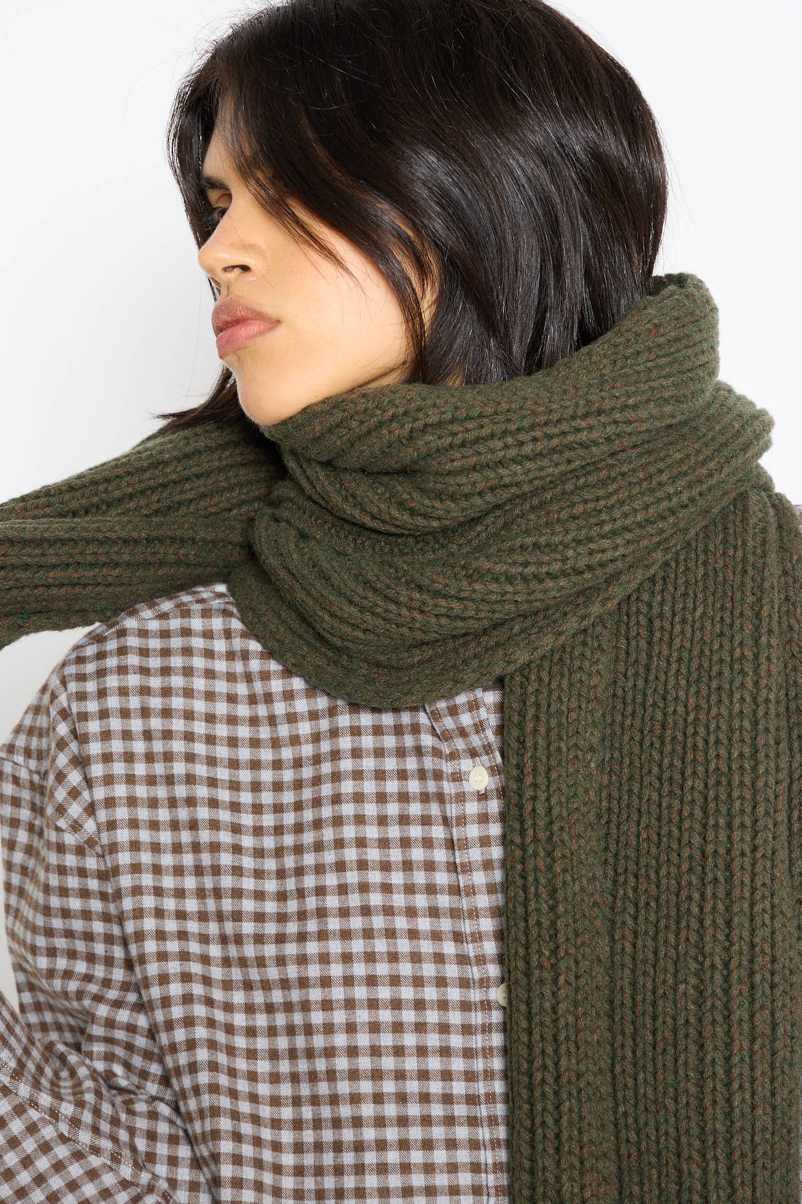 A person with medium-length dark hair, wearing a chunky knit Mea Scarf in Foret by Baserange made from a recycled wool blend and a brown checkered shirt, is looking to the side.